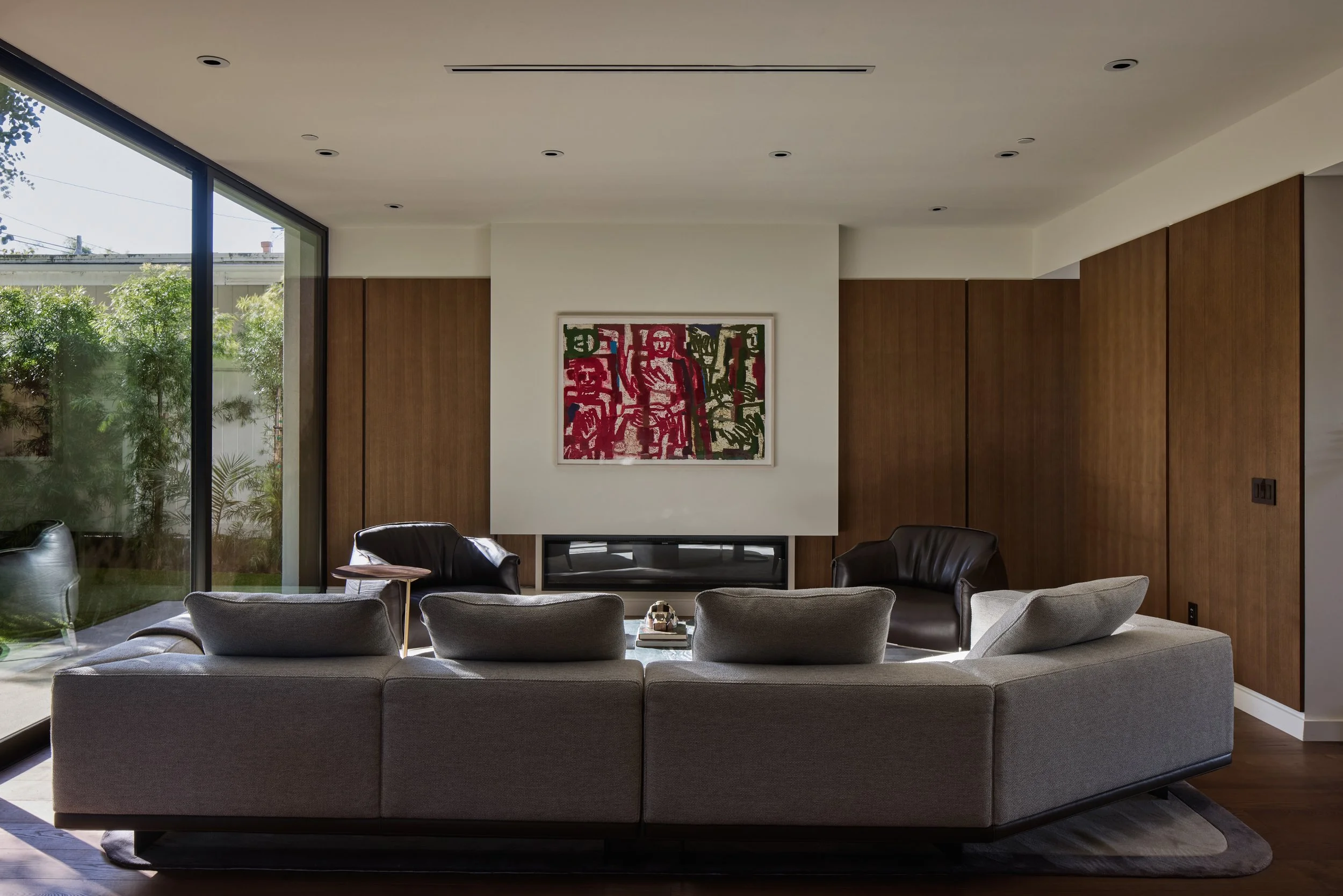 A modern living room with a large gray sofa, two armchairs, and a rectangular coffee table. There are large sliding glass doors on the left side that let in natural light and show greenery outside. The wall behind the sofa has wooden paneling and art