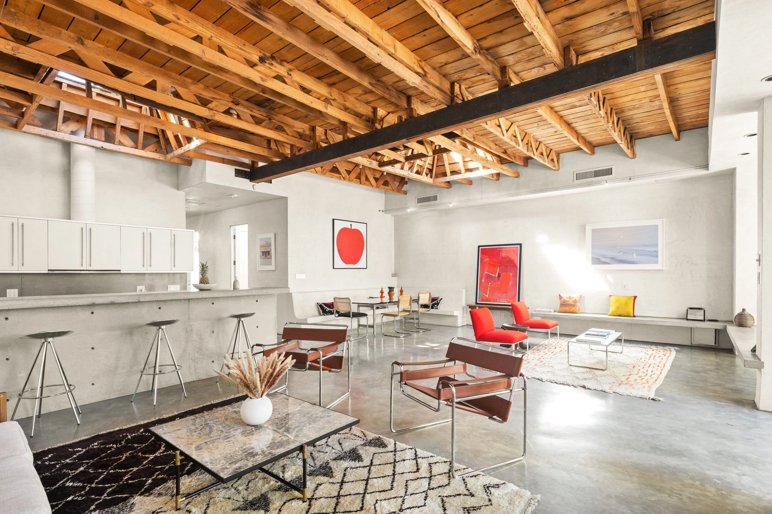 A spacious, modern living room with an open floor plan, featuring a high wooden ceiling with exposed beams, white walls with artwork, a kitchen area with white cabinets and a marble countertop, colorful furniture including red chairs, a light-colored