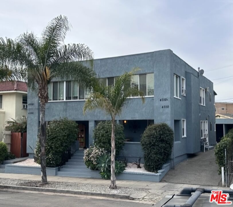 A two-story blue residential building with a flat roof, surrounded by palm trees and bushes, with a small front porch area and stairs leading up to the entrance, located on a city street.