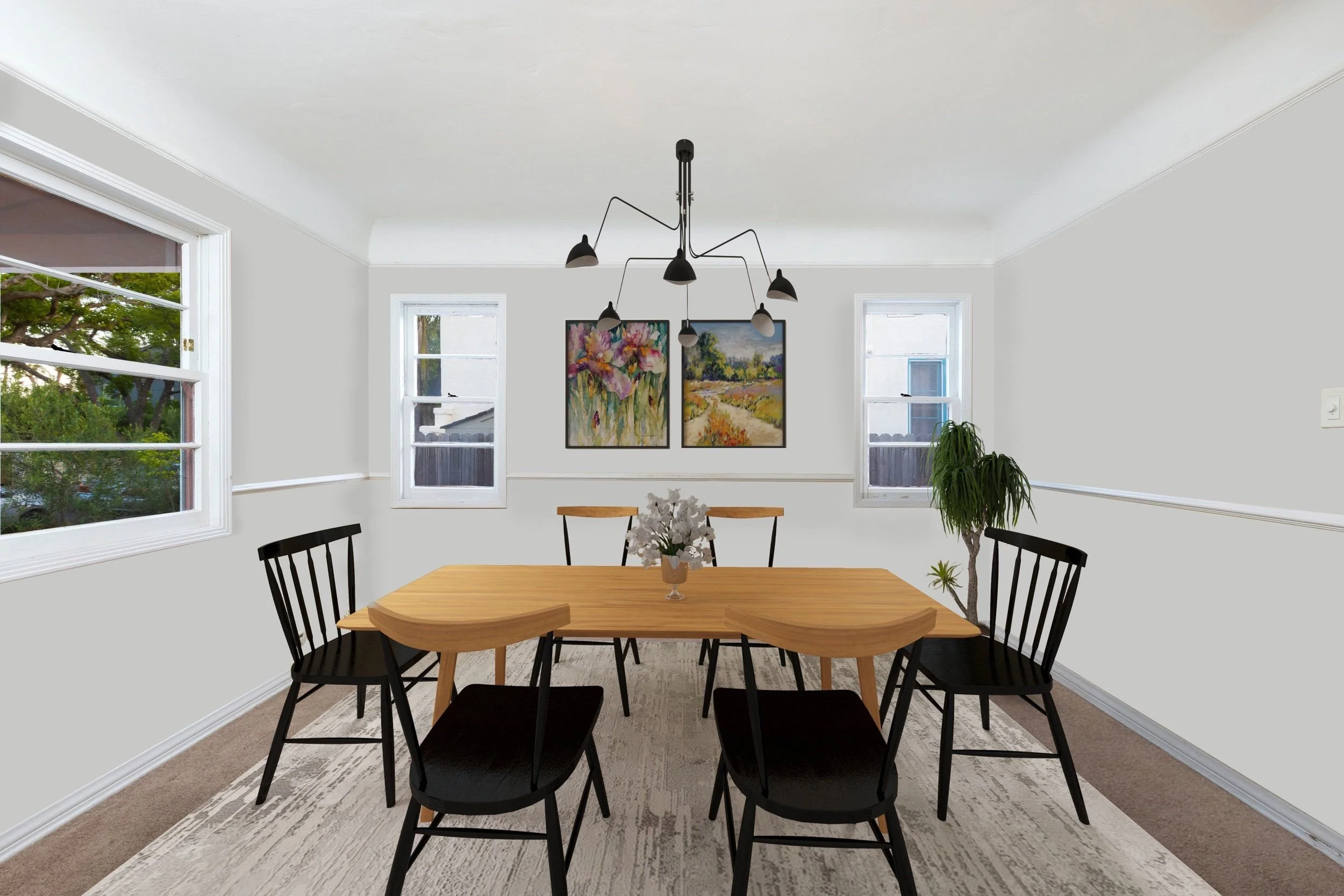 A dining room with a wooden table, six black chairs, a vase with white flowers, large windows with greenery outside, and artwork on the wall.