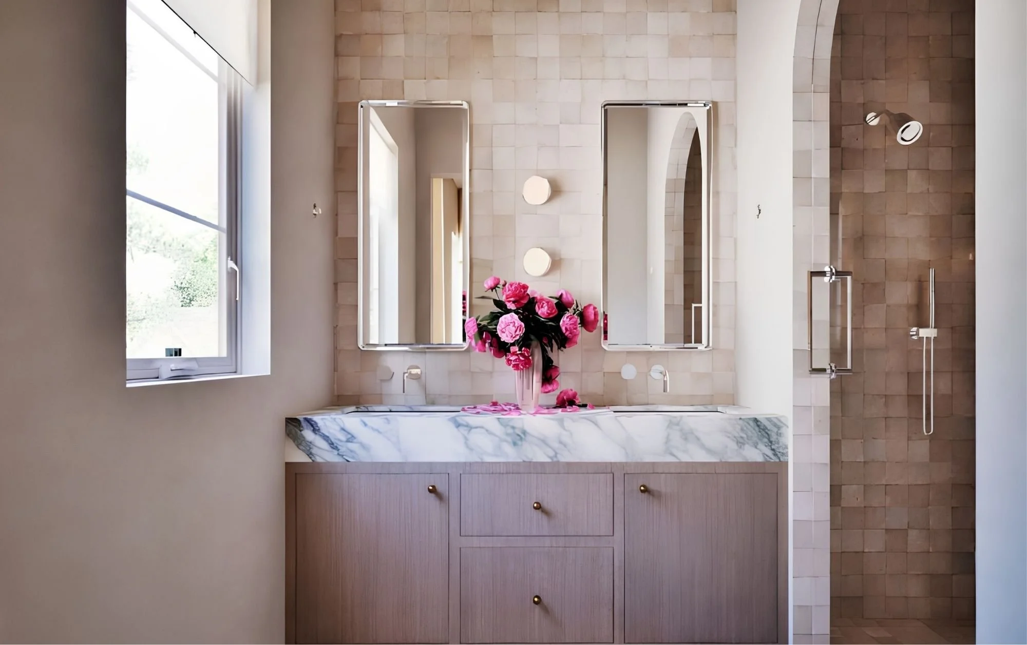 A modern bathroom with two rectangular mirrors above a marble countertop with a vase of pink flowers, beige tiled walls, a window on the left, and a shower area on the right.