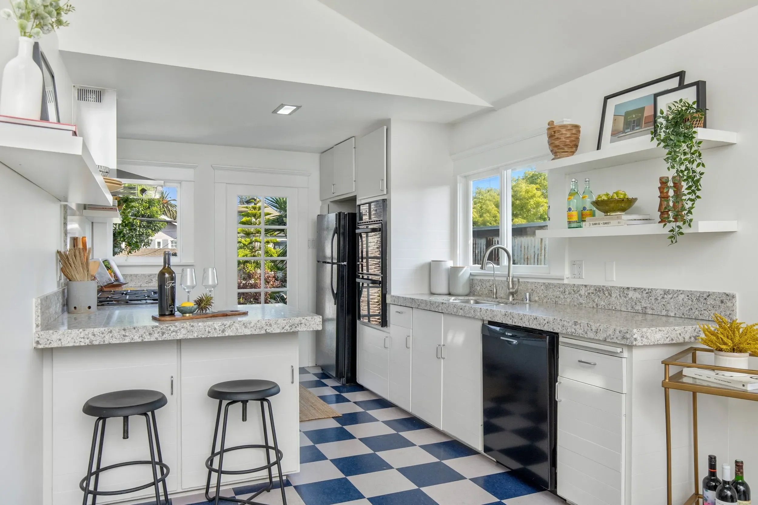 A bright kitchen with white cabinets, granite countertops, and a black refrigerator. There are open shelves with framed artwork and decorative items. Two black barstools are positioned at a breakfast counter. The floor has a blue and white checkered 