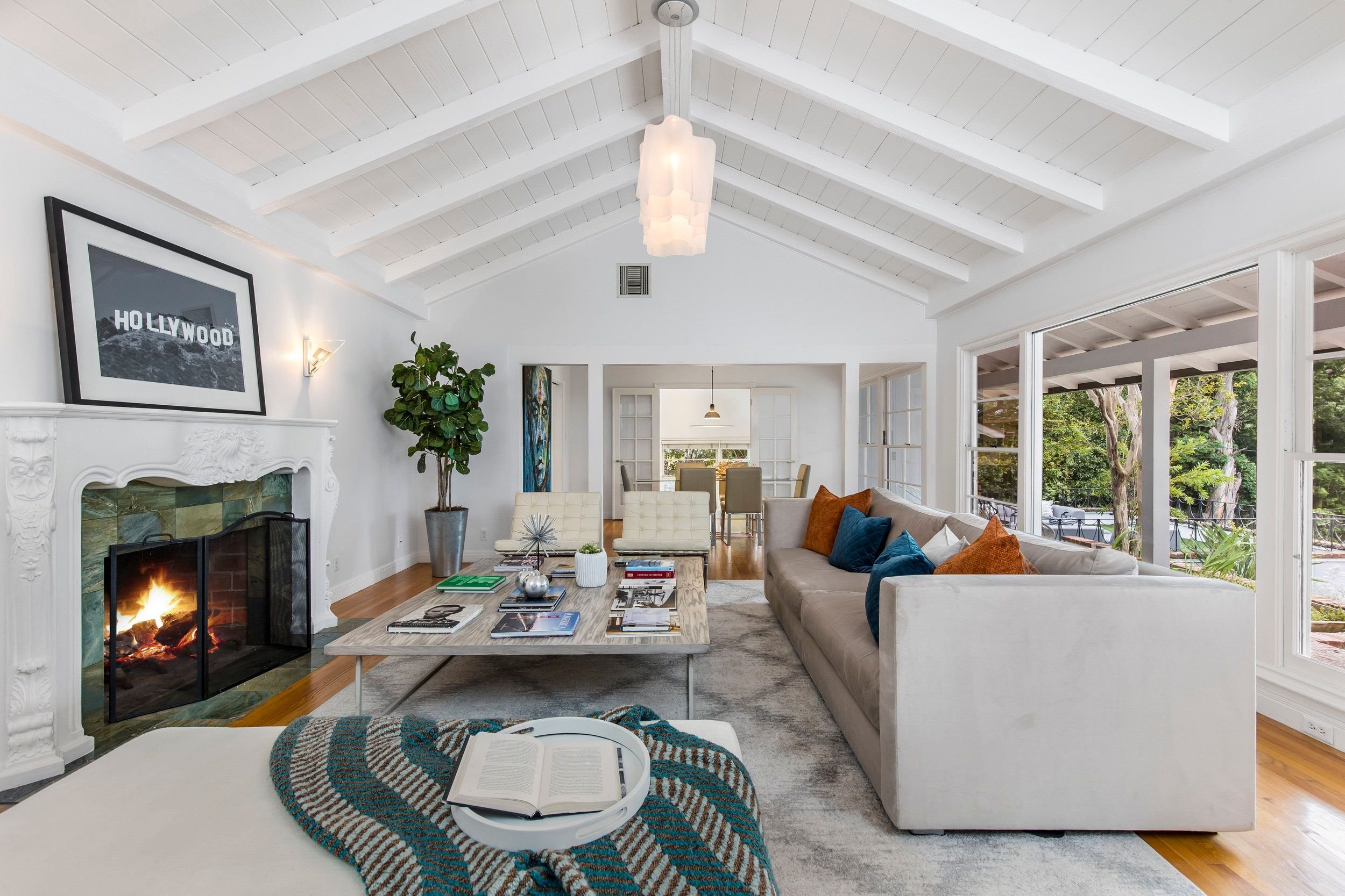 Bright living room with white walls and high vaulted ceiling with exposed beams, large windows overlooking green trees, a fireplace with white ornate mantel, a beige sofa with colorful pillows, a coffee table with magazines, and an open dining area i