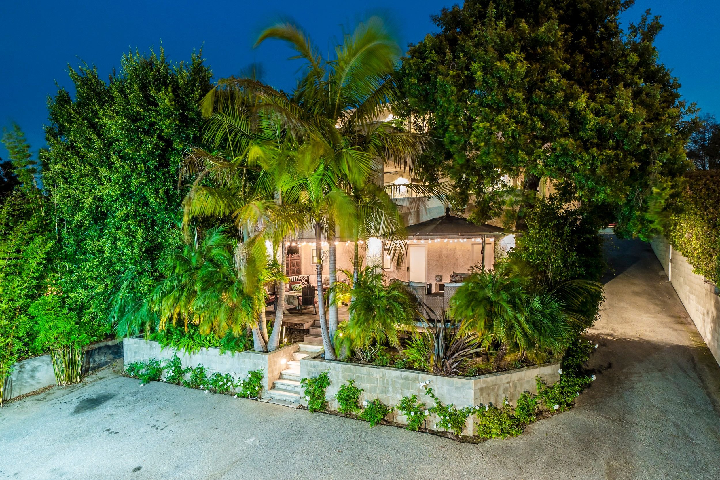 Nighttime view of a backyard garden with lush green trees, a patio area with outdoor furniture, string lights, and colorful outdoor lighting illuminating the scene.