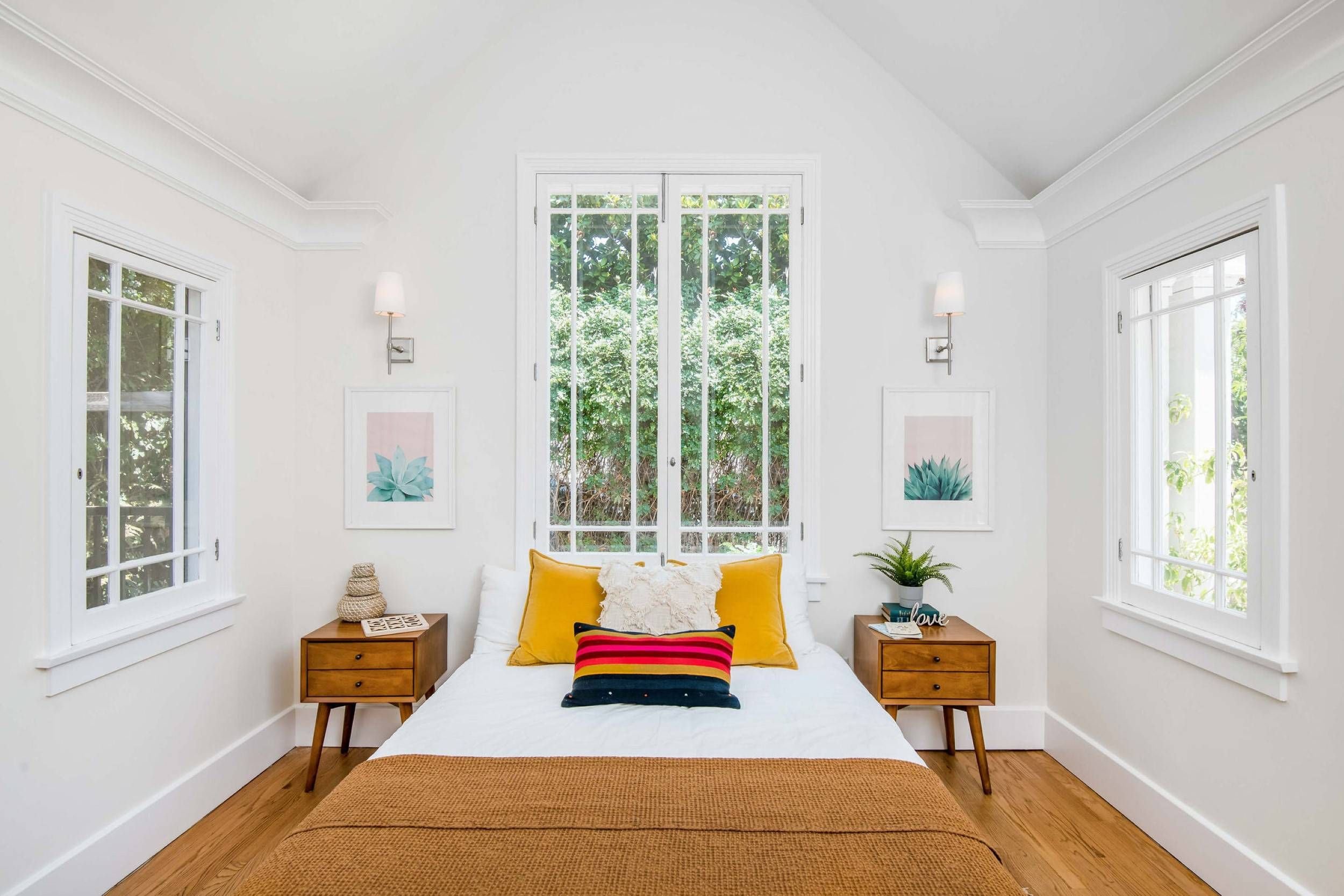 Bright bedroom with white walls, large windows, and outdoor greenery view. Wooden nightstands on each side of the bed with lamps, artwork, and a potted plant.