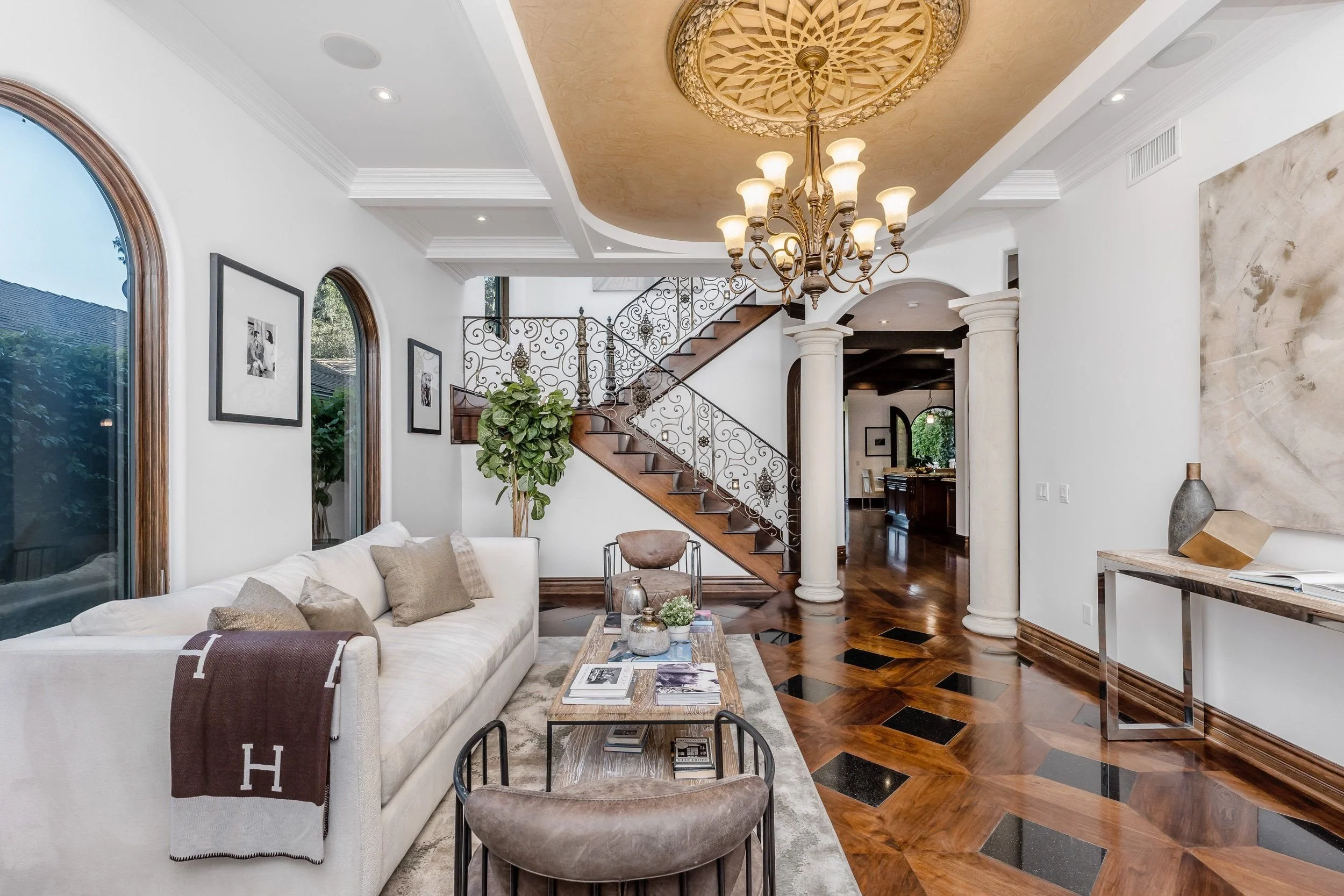 Living room with white sofa, wooden coffee table, artwork on the walls, a staircase with ornate railing, chandelier, and wooden flooring.