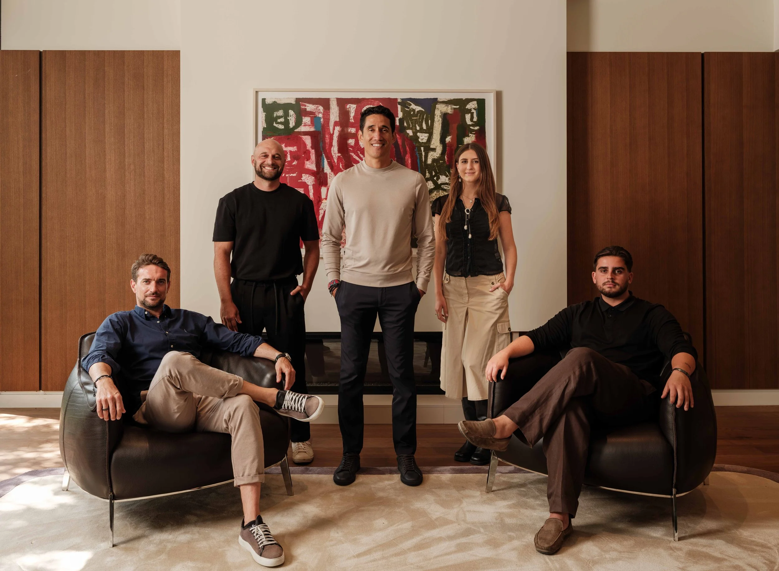 Group of six diverse young adults posing in a modern living room, with one sitting on a couch and five standing, in front of an abstract painting on a white wall.