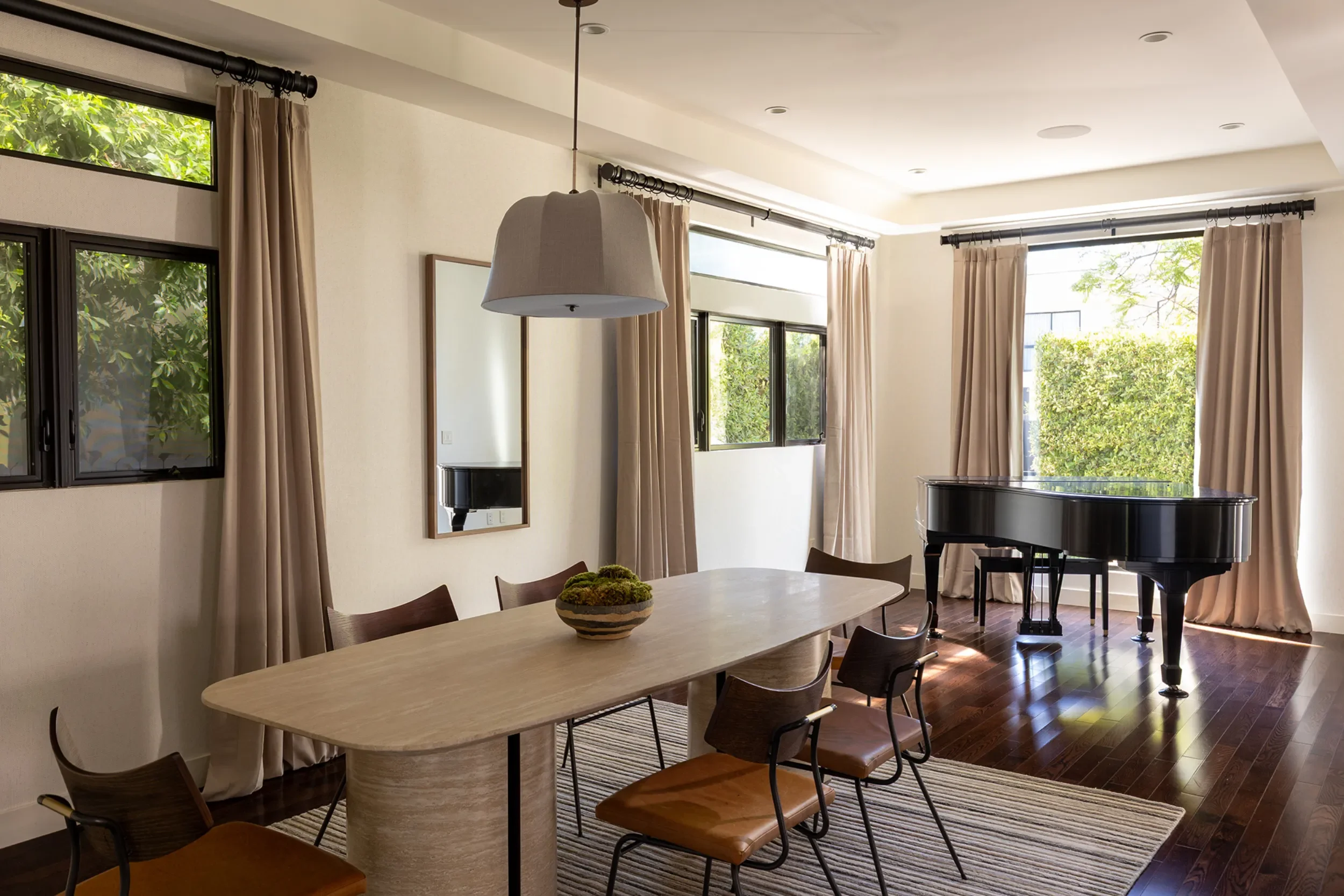 A room with large windows and beige curtains, a wooden dining table with chairs, a decorative bowl with moss, a mirror on the wall, a grand piano, and a lampshade hanging from the ceiling.