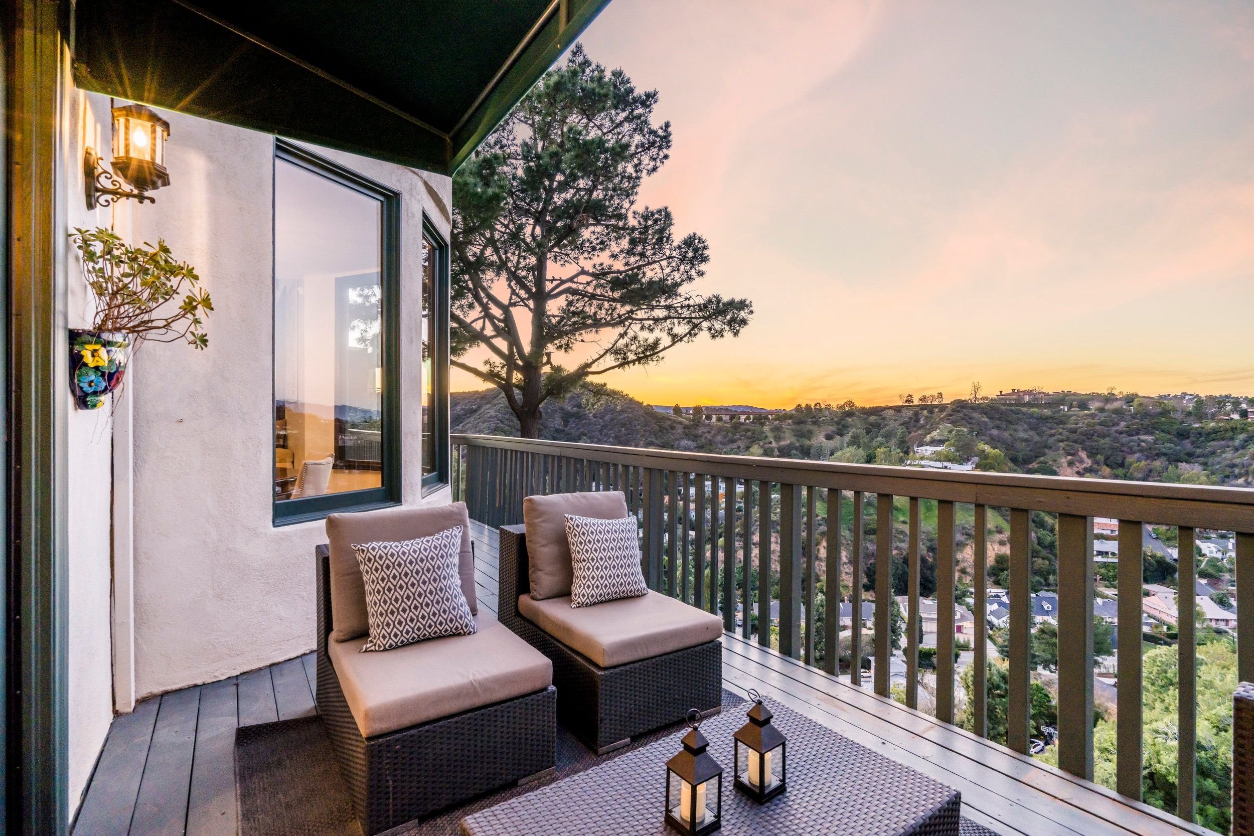 Balcony with two cushioned chairs, decorative pillows, wooden floor, lanterns, and a view of rolling hills and trees at sunset.