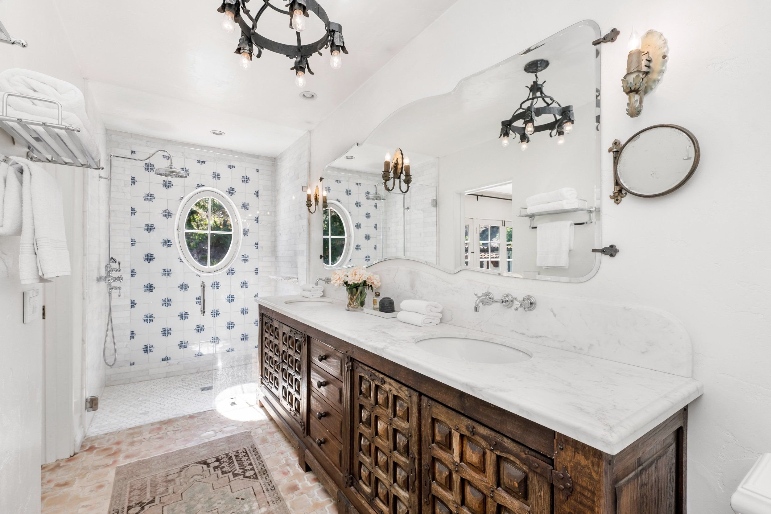 A bathroom with a large mirror above a rustic wooden vanity with a marble countertop. There are white towels on a rack and shelves, vintage light fixtures, a decorative lamp, and an oval window with greenery visible outside. The shower area has patte