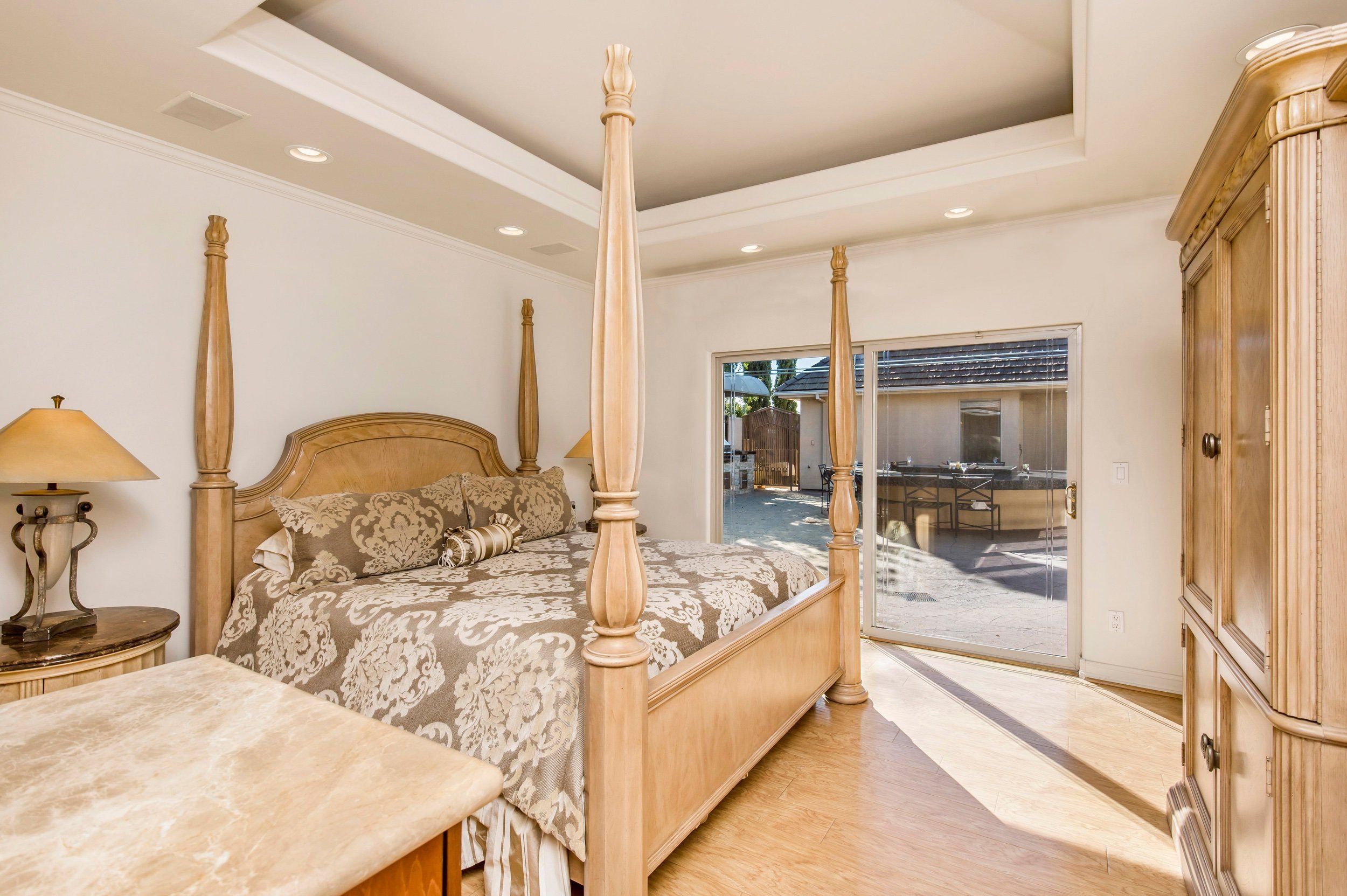 A bedroom with a four-poster wooden bed with beige and cream patterned bedding, a nightstand with a lamp, and large sliding glass doors leading to an outdoor patio area.