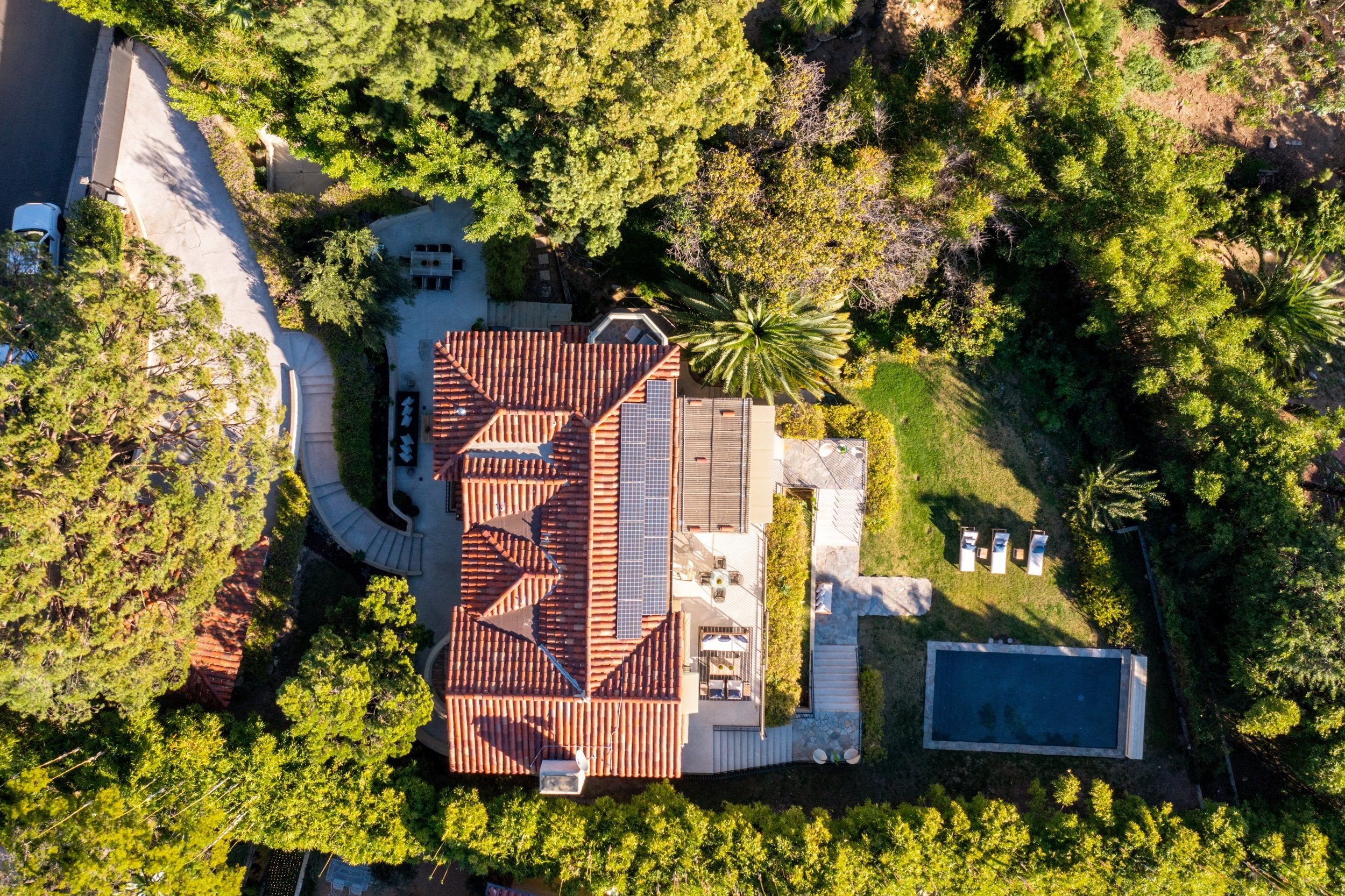 Aerial view of a house with a red tiled roof surrounded by dense green trees and a lush backyard with a swimming pool.