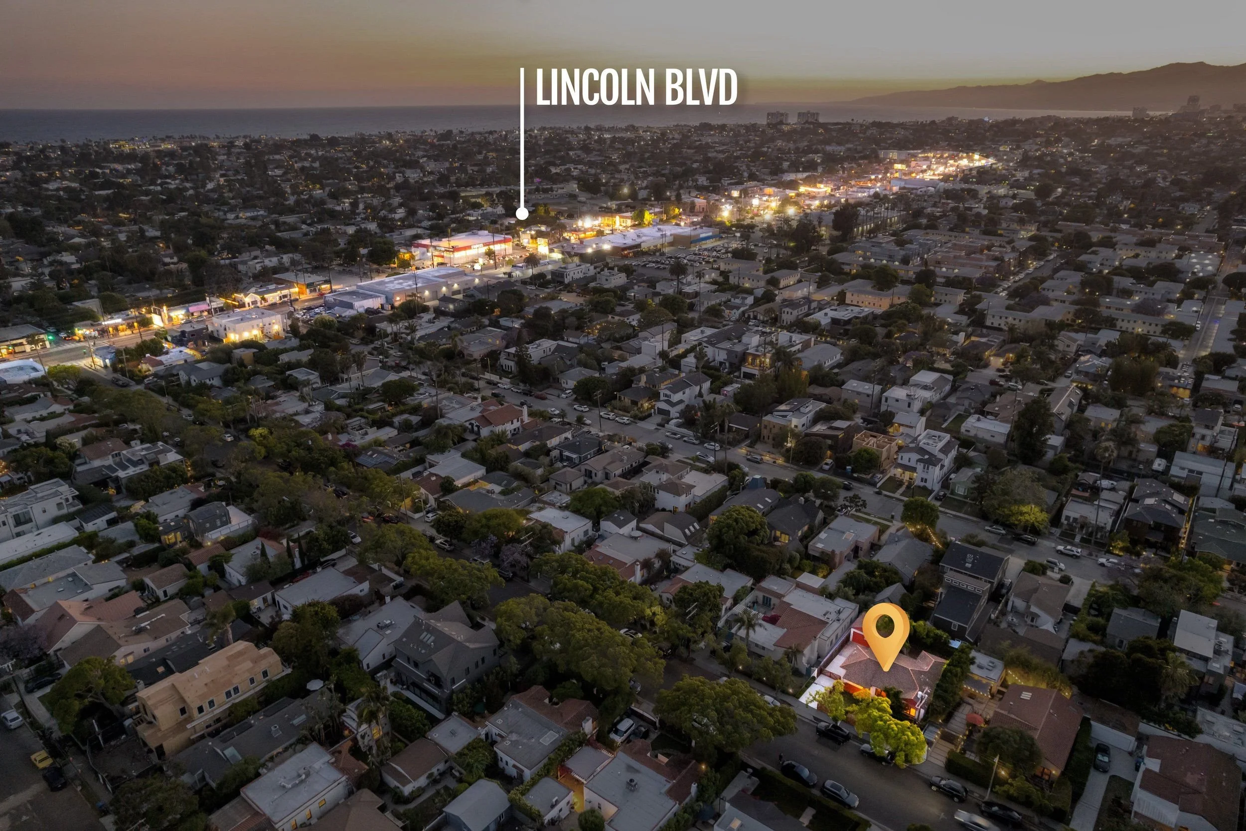 An aerial view of a cityscape at dusk, with residential houses, commercial buildings, and streets illuminated by streetlights. The horizon shows the ocean and mountains in the distance, with the text 'Lincoln Blvd' pointing to a location in the city.