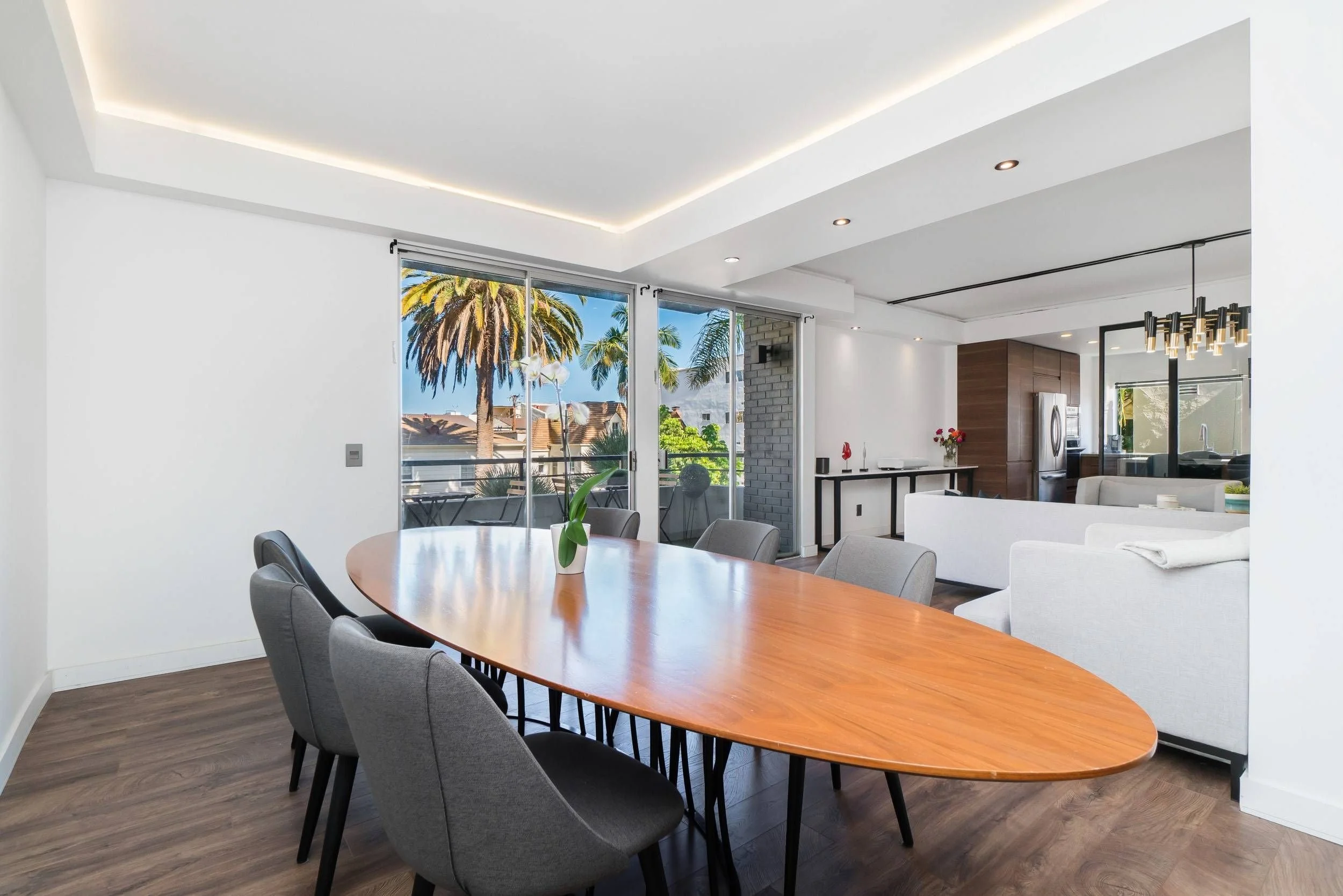 Modern dining and living area with large glass doors opening to a balcony with palm trees, featuring a wooden dining table, gray upholstered chairs, white sectional sofa, and ceiling lighting.