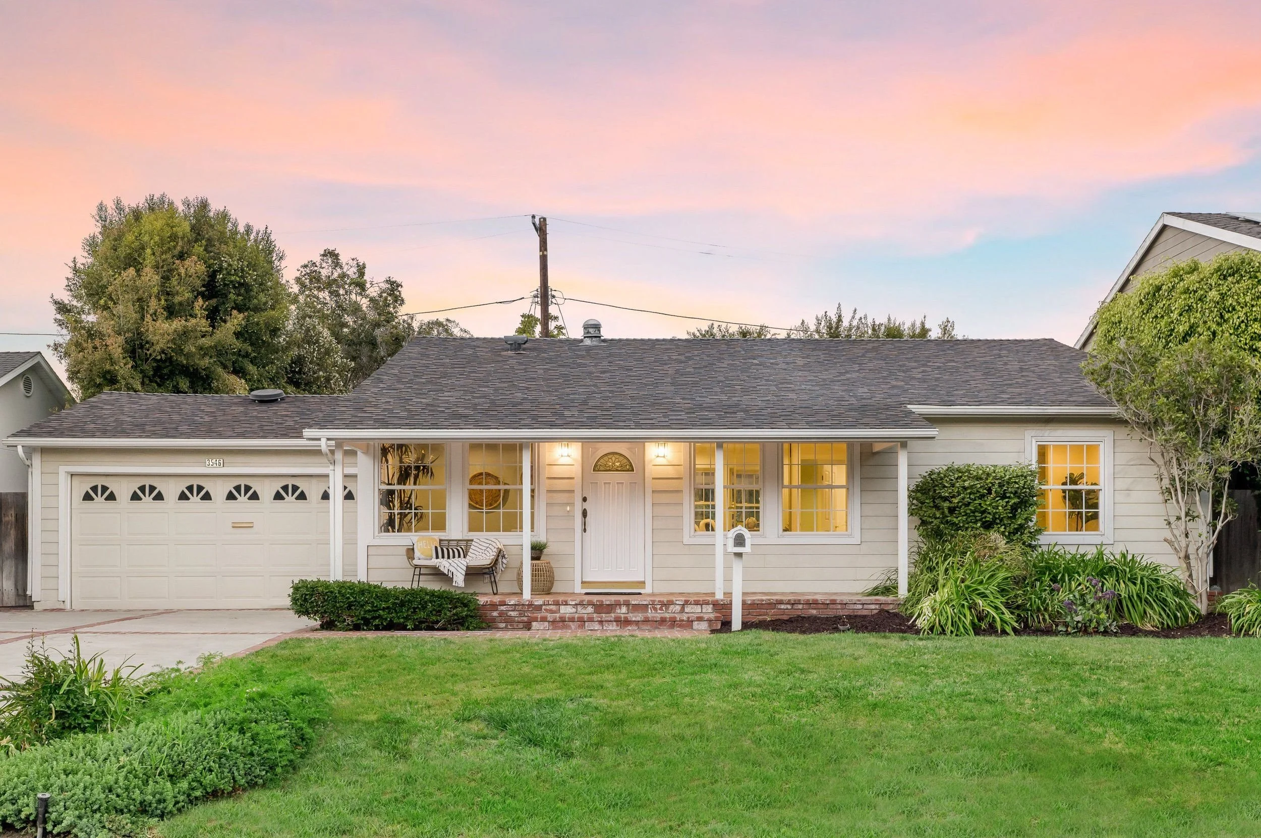 A cozy white house with a gray shingle roof, front porch, and well-maintained garden, illuminated by warm interior lights, set against a sky with pink and blue clouds.