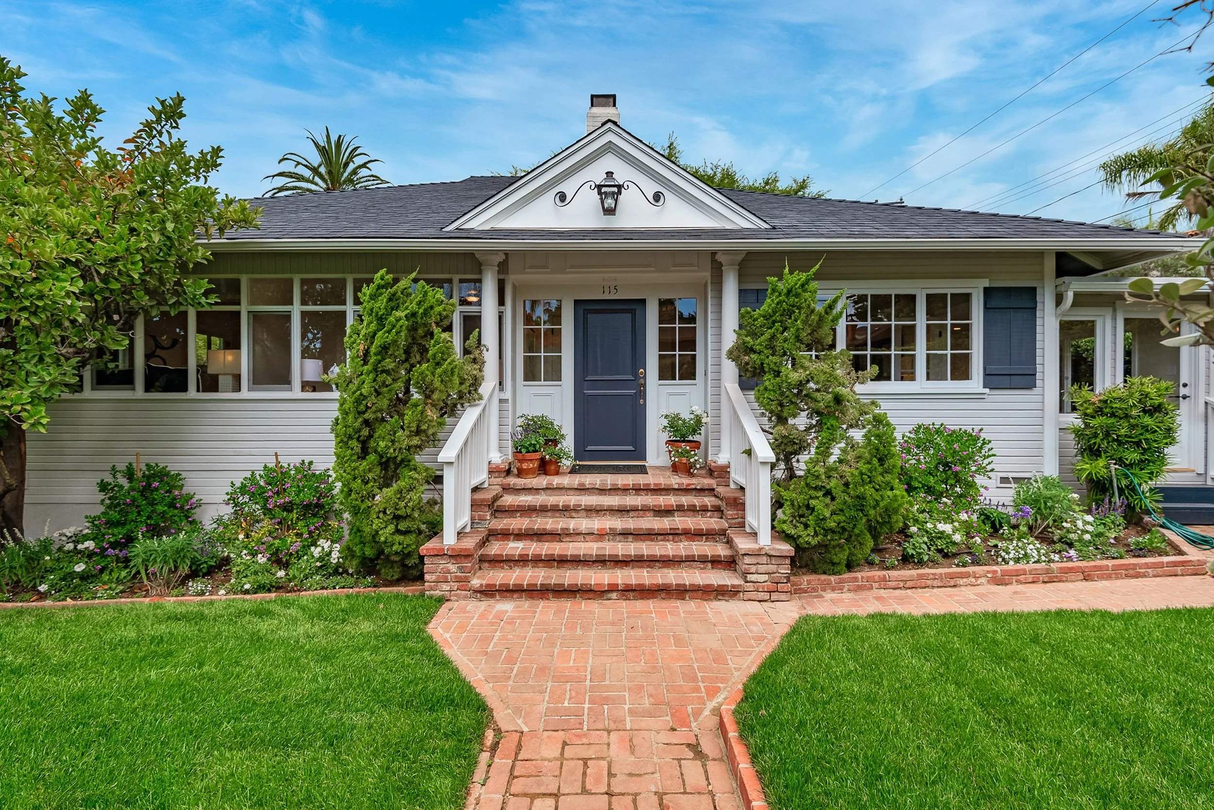 Front view of a white house with a black door, brick steps, a brick pathway, lush green lawn, and various shrubs and flowers in the front yard. The house has large windows and a gabled roof with a lantern hanging under the gable.