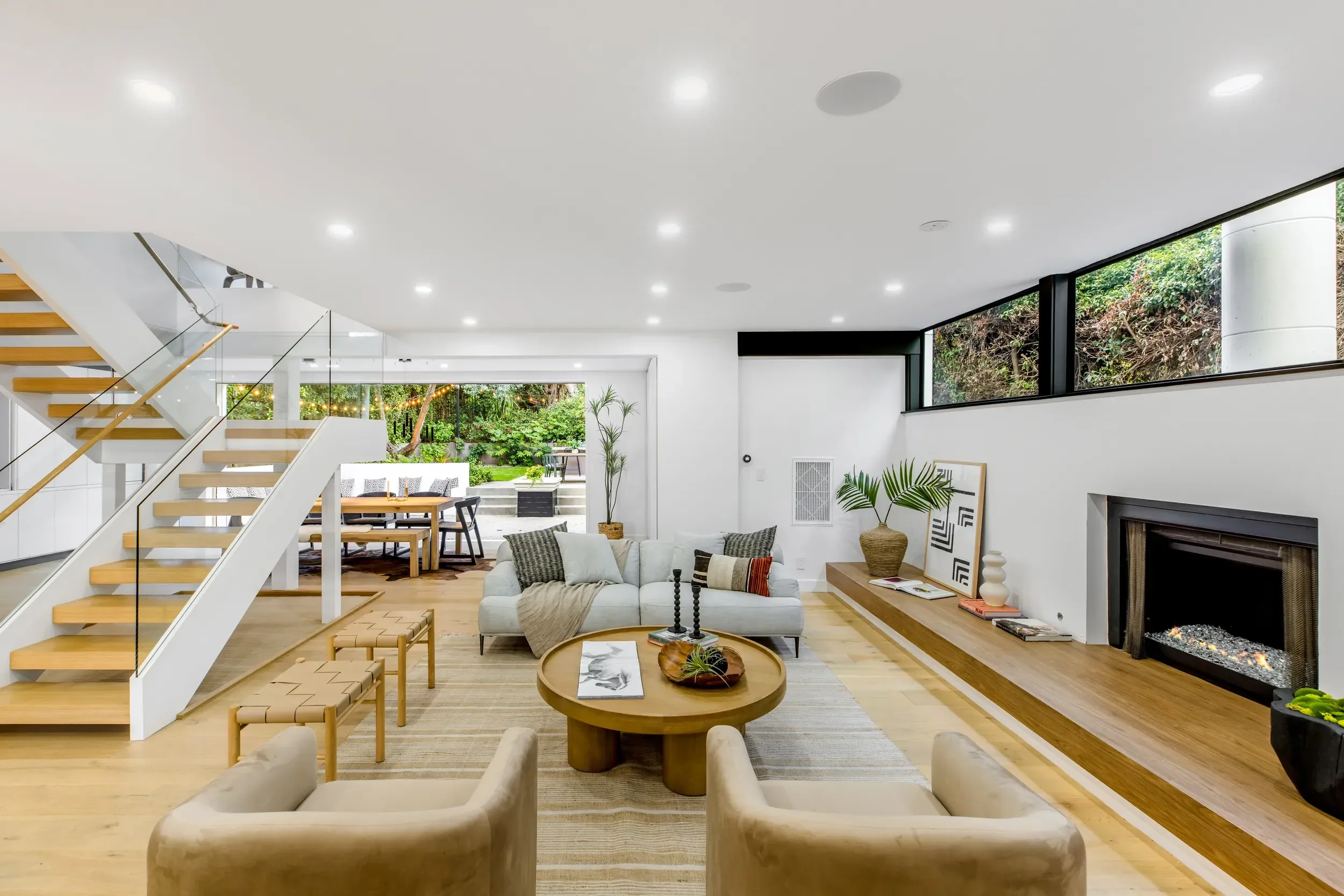 Modern living room featuring a white sofa, wooden coffee table, fireplace, large windows overlooking greenery, and a staircase with wooden steps and glass railing.