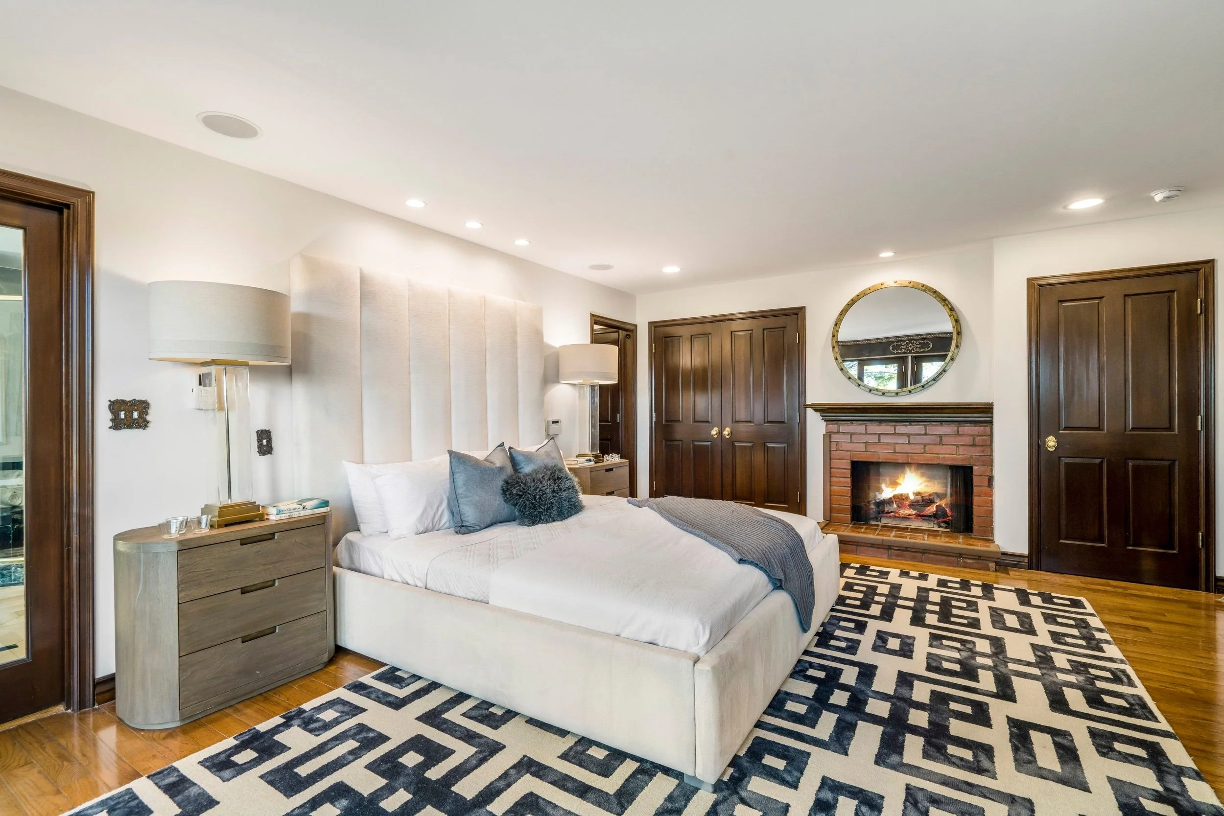 A bedroom with a white bed, gray pillows, a wooden nightstand, and a fireplace with a mirror above it, wood-paneled doors, and a geometric rug.