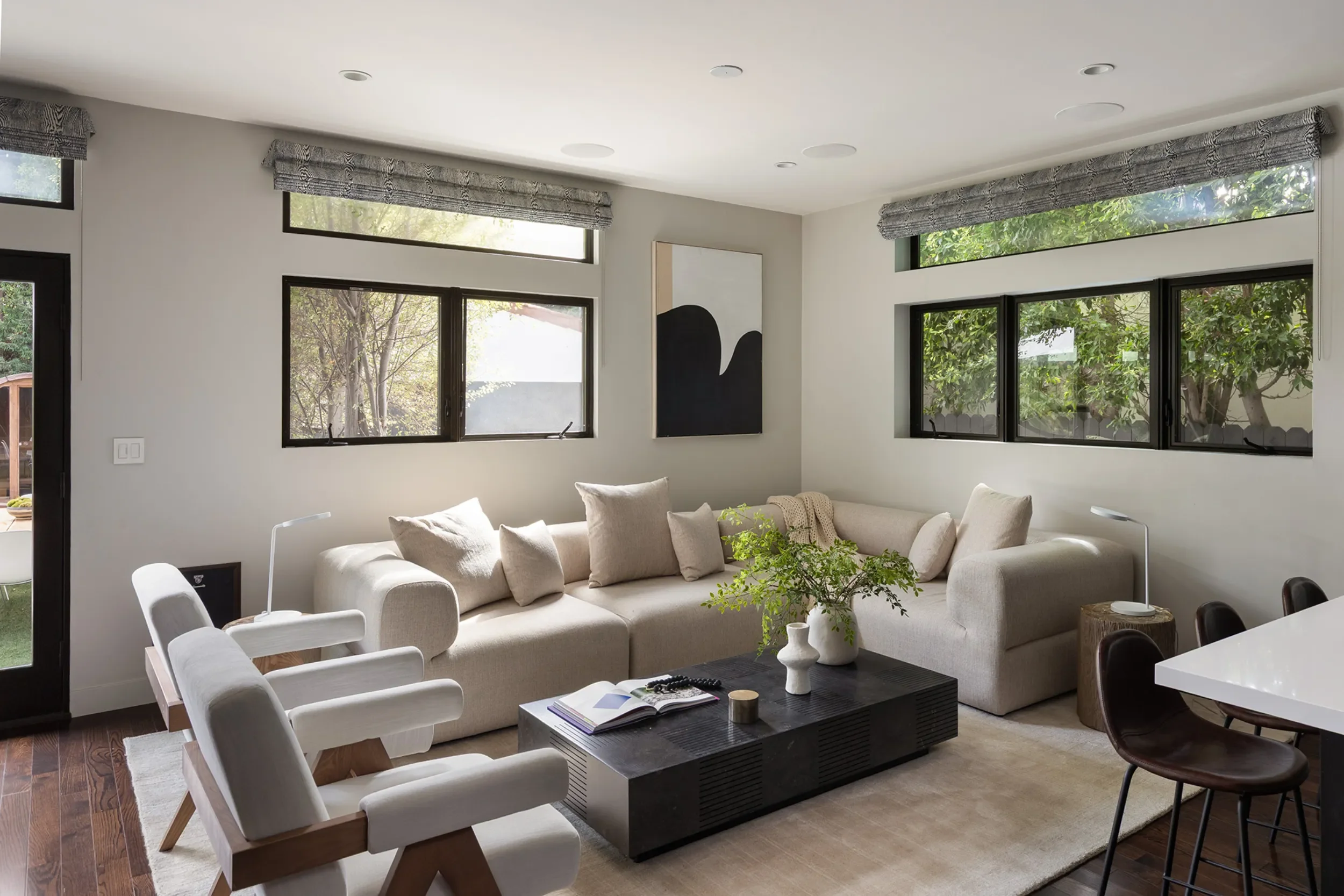 Living room with beige sofa, black coffee table with books and decor, white armchairs, large windows with gray valances, and decorated with a black abstract painting.