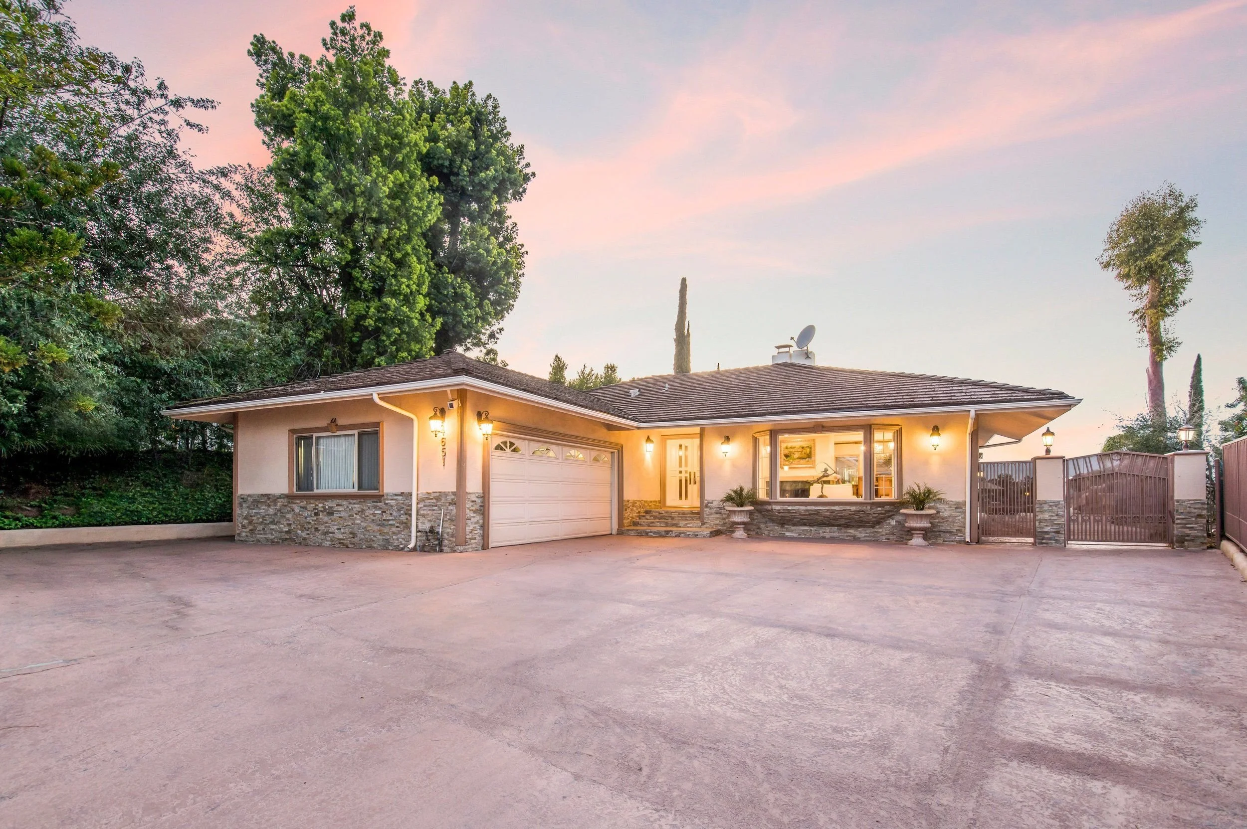 A single-story house with a garage door, front yard, and outdoor lights. The house has a tiled roof, stone accents, and large windows. Tall trees surround the house, and the sky is pink and purple indicating sunset or dawn.