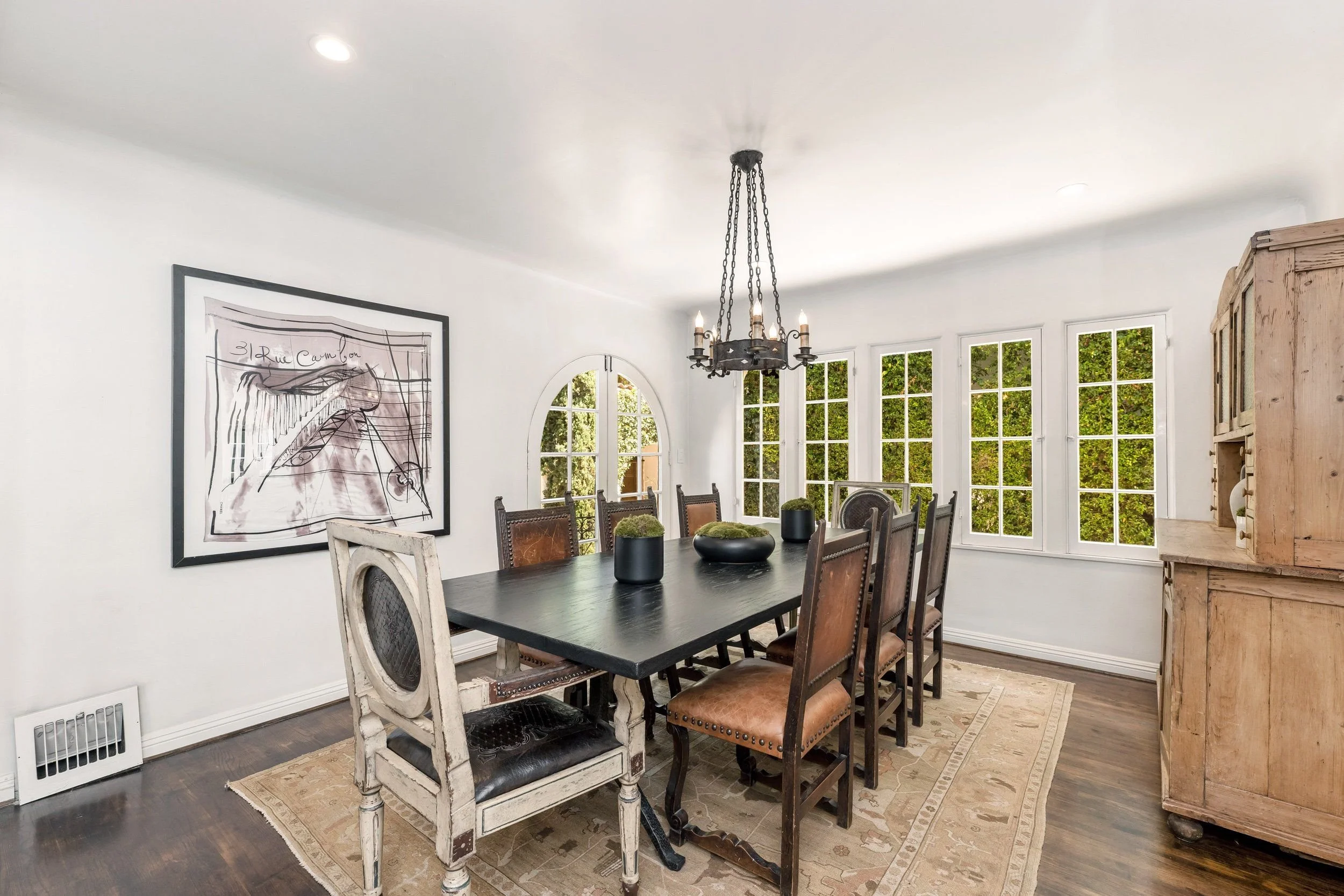 A dining room with a large dark wood table and various chairs, a chandelier hanging from the ceiling, large windows with greenery outside, a framed artwork on the wall, a wooden cabinet on the right, and a decorative rug under the table.