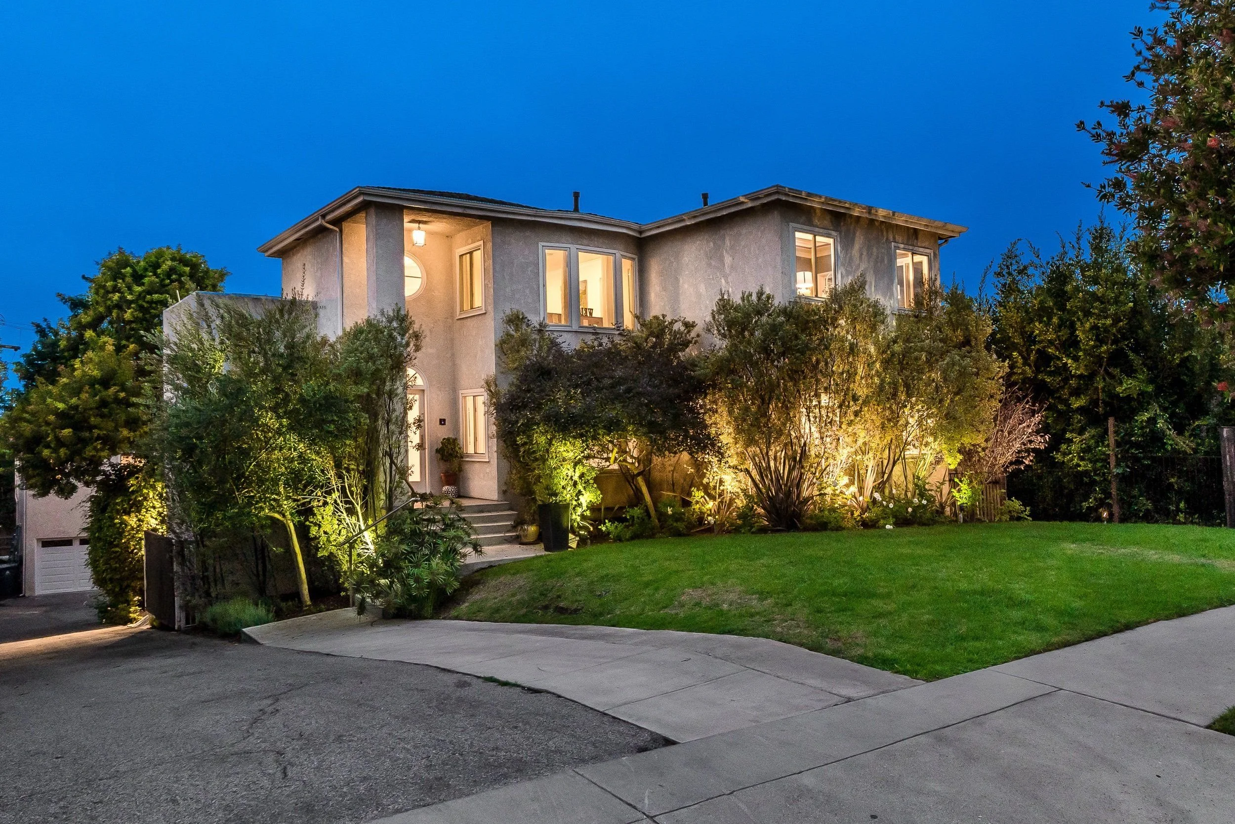 A two-story house with large windows, illuminated from inside, surrounded by trees and shrubs at dusk. A driveway leads up to the house with a lawn in front.