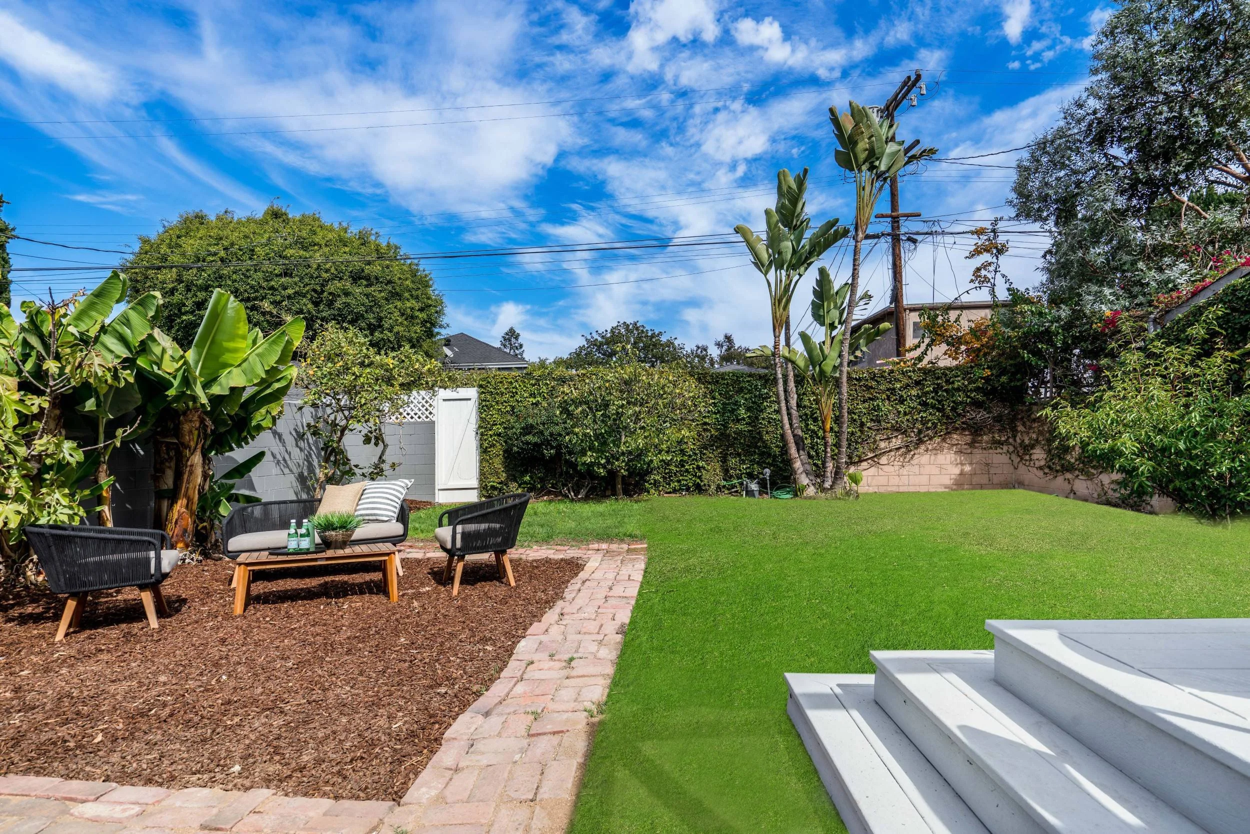 A backyard garden with a patio area, black outdoor chairs, a small table with drinks, trimmed grass, trees and plants, a white fence, and electrical wires against a blue sky with clouds.