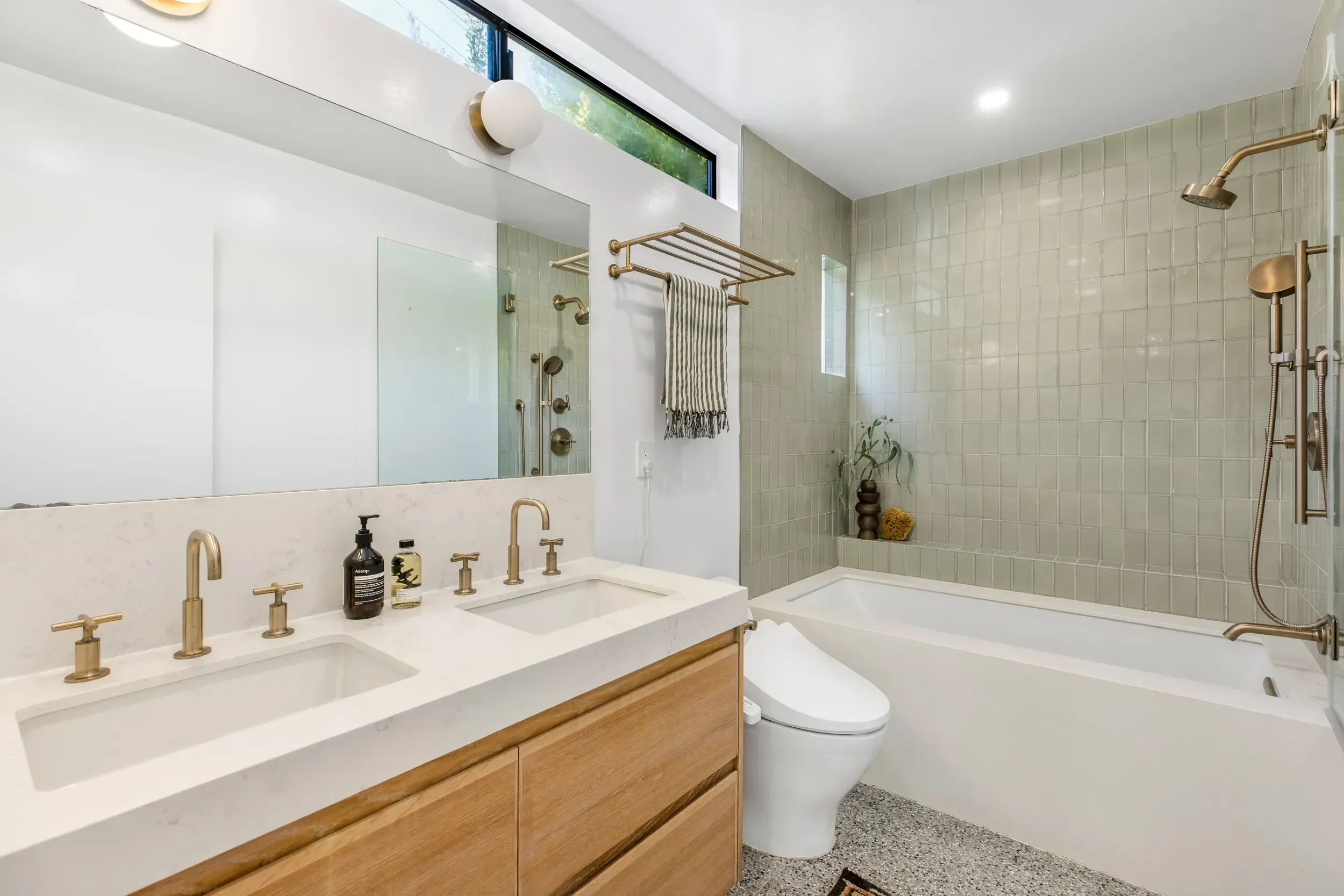 Modern bathroom with a dual-sink vanity with gold fixtures, a large mirror, a bidet, a bathtub with a shower, beige tiles in the shower area, and a narrow horizontal window near the ceiling