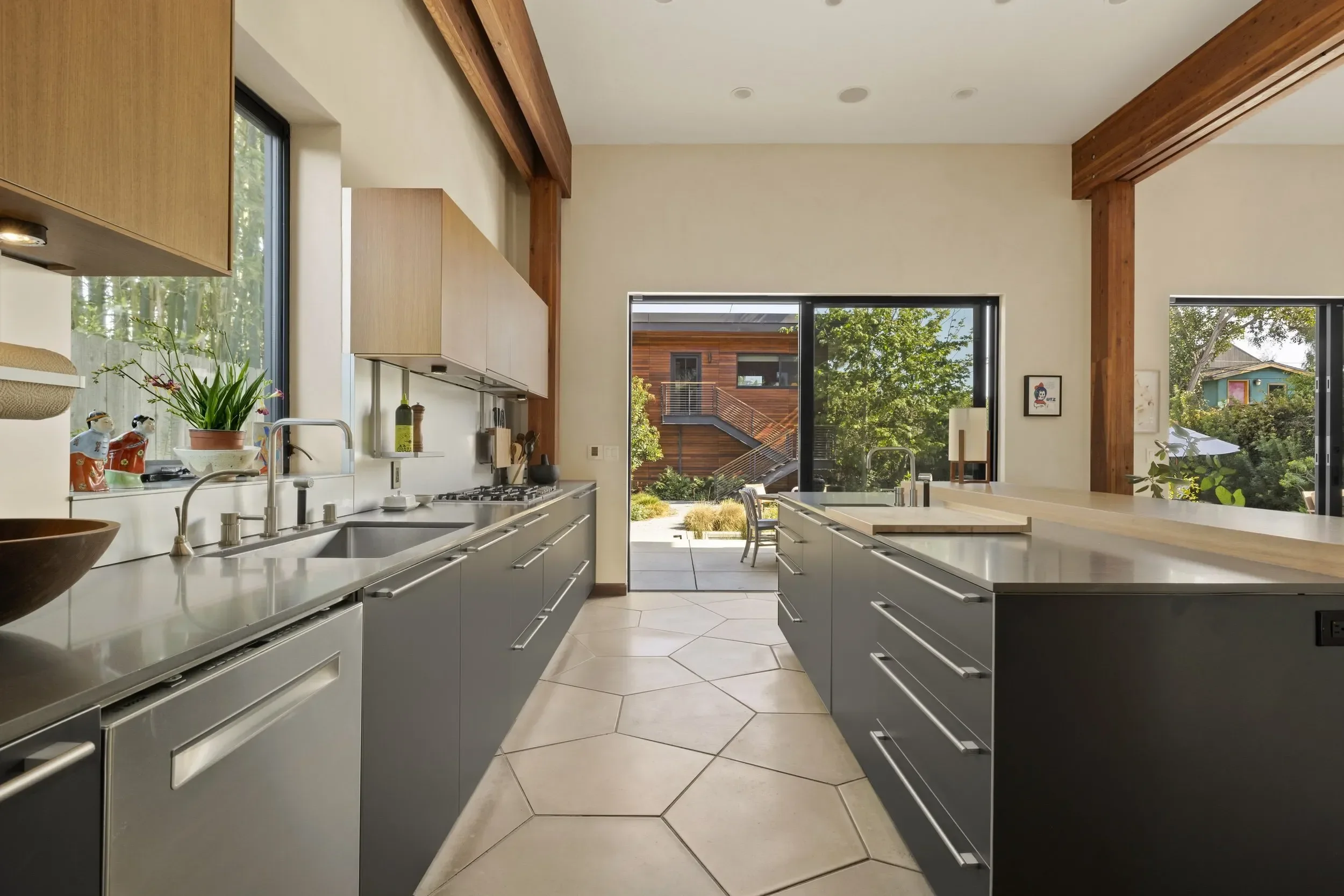 Modern kitchen with gray cabinets, white countertops, and large windows showing outdoor greenery and neighboring buildings with wooden and blue exterior.
