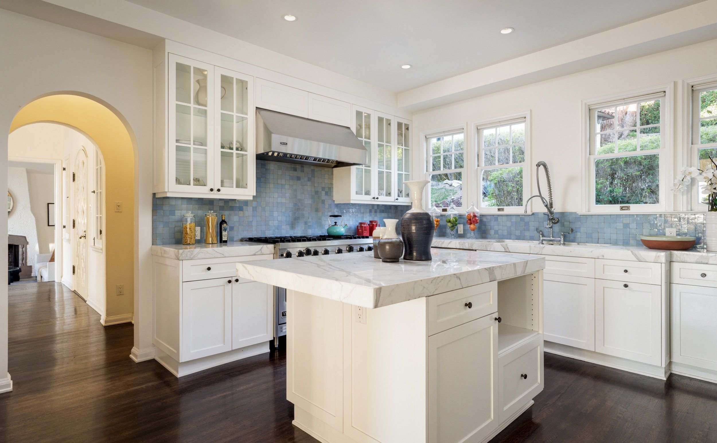 A bright kitchen with white cabinets, a marble countertop, a blue tile backsplash, large windows, and decorative vases and food containers on the counter.