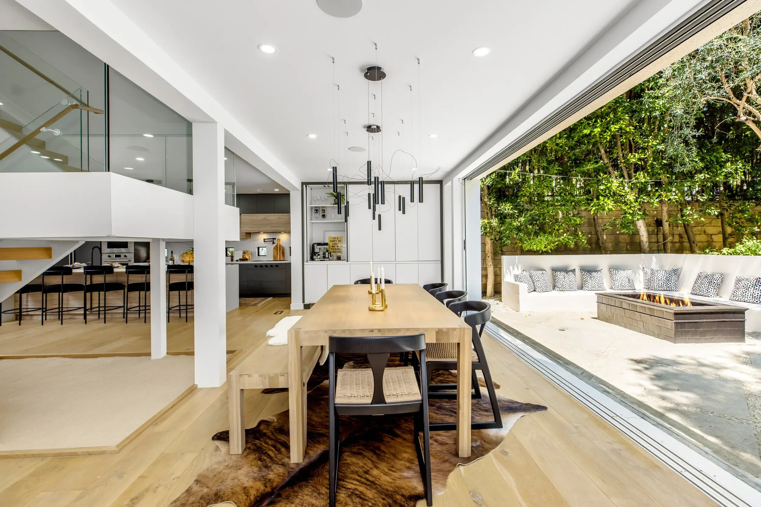 Modern dining area with a large wooden table, black chairs, and a spacious open-plan kitchen in the background. Large sliding glass doors open to an outdoor patio with a built-in fire pit surrounded by greenery.