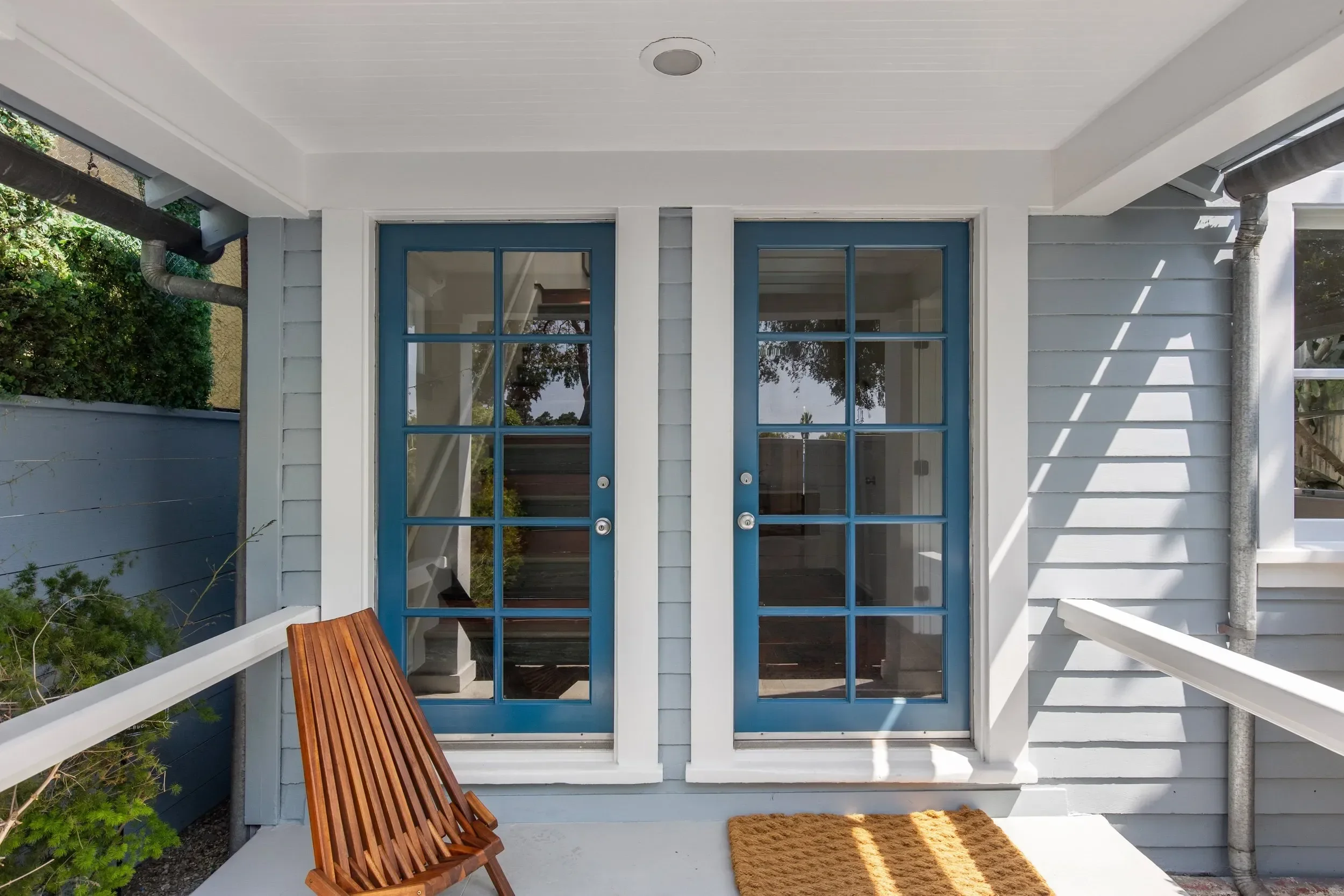 Front porch of a house with a white ceiling, blue-framed glass doors, a wooden bench, a brown doormat, and part of a gray painted wall with a drainpipe, surrounded by greenery.
