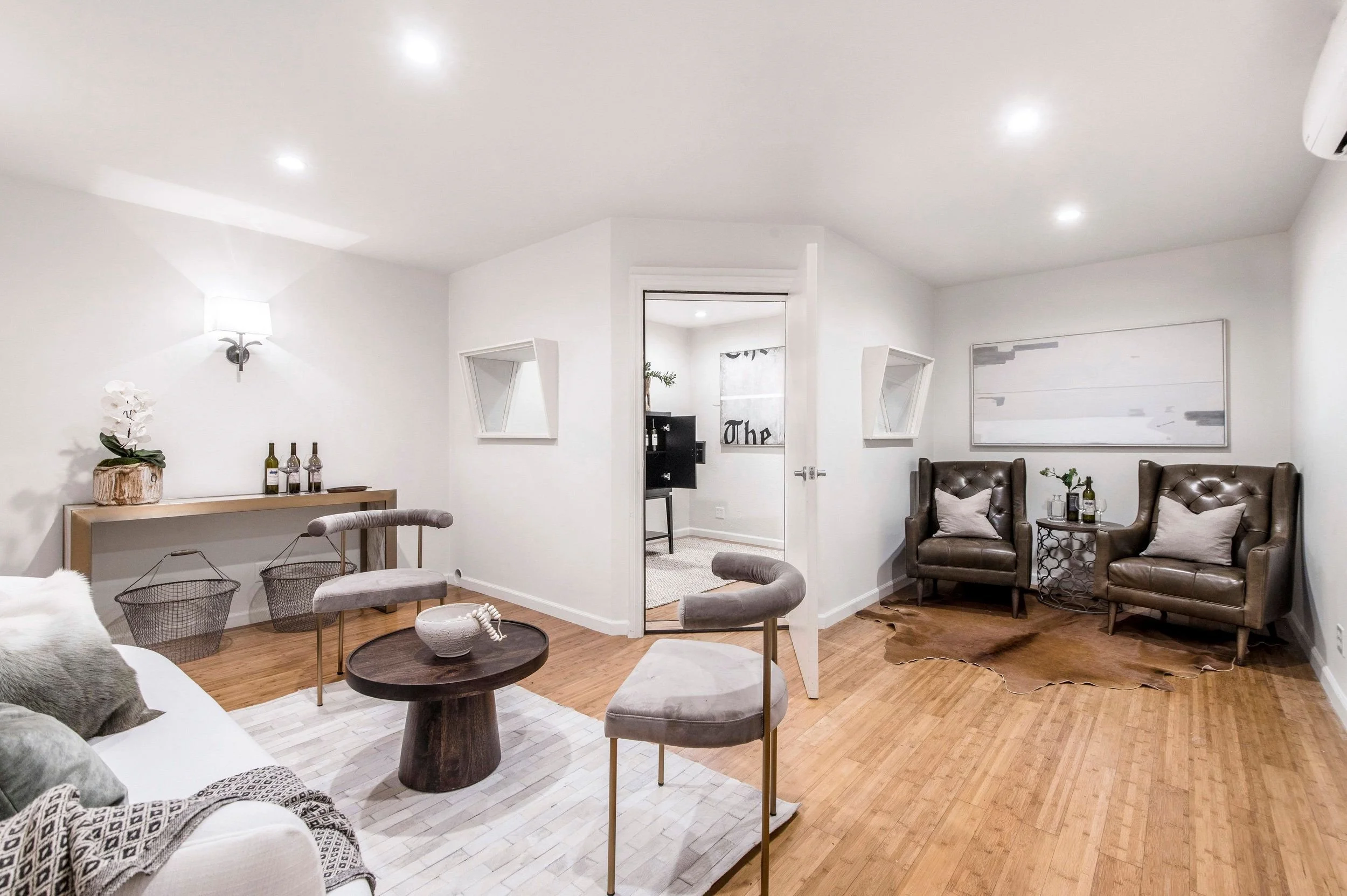 Living room with white walls, wooden floor, and modern furniture including brown upholstered chairs, a white sofa, and decorative items like potted plants and bottles.