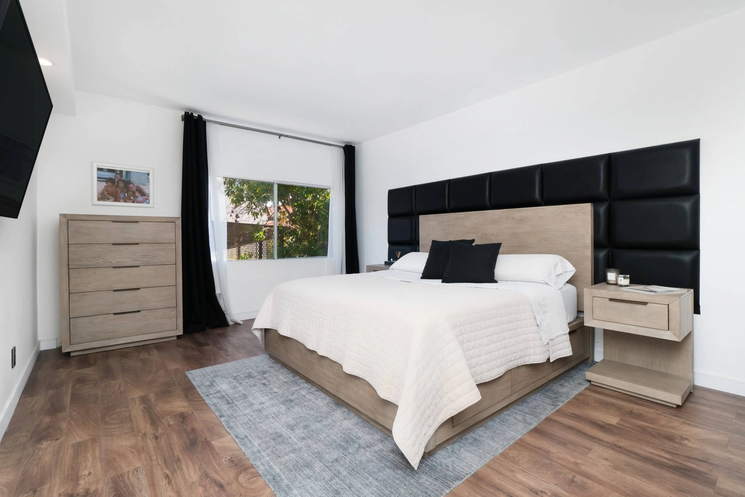 A modern bedroom with a large bed, black and white bedding, wooden nightstands, a dresser, window with black and white curtains, a wall picture, and a TV mounted on the wall.