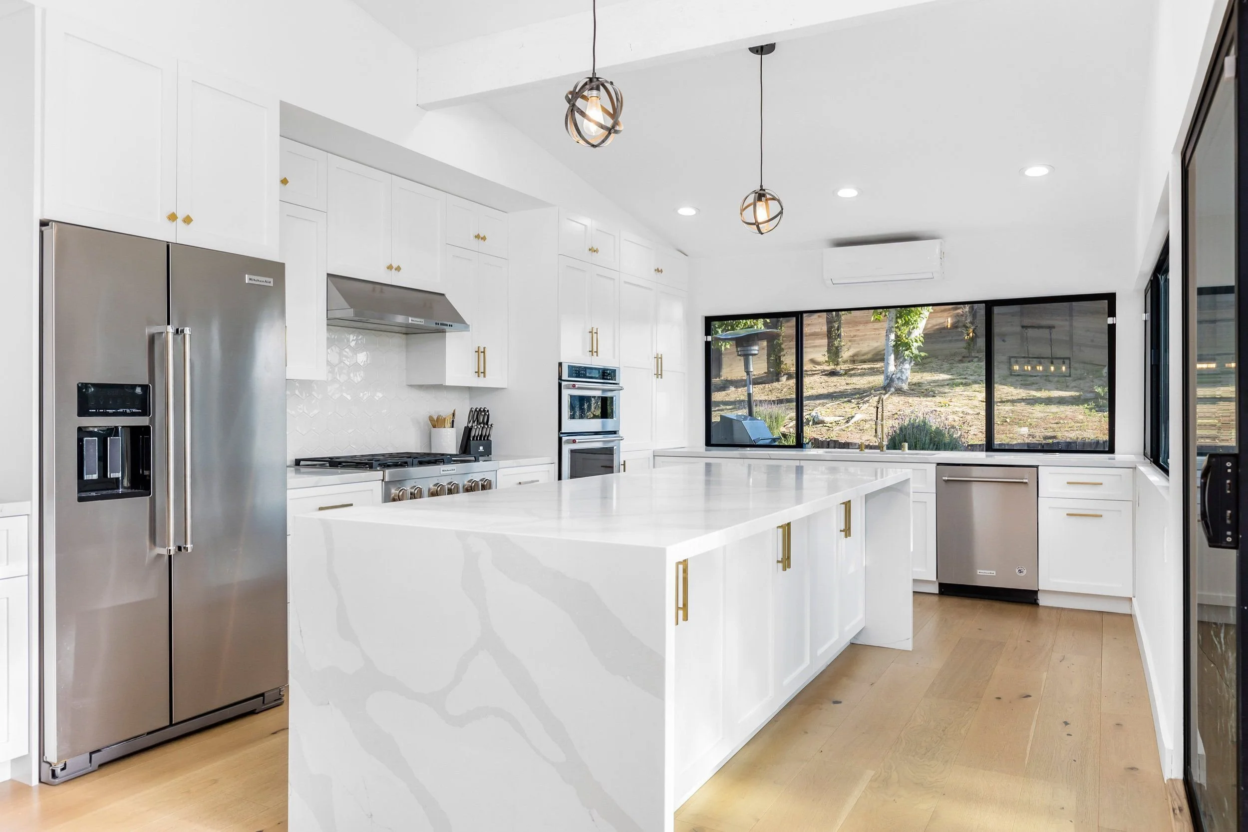 Modern white kitchen with large island, stainless steel refrigerator, built-in oven, and large windows showing outdoor greenery.