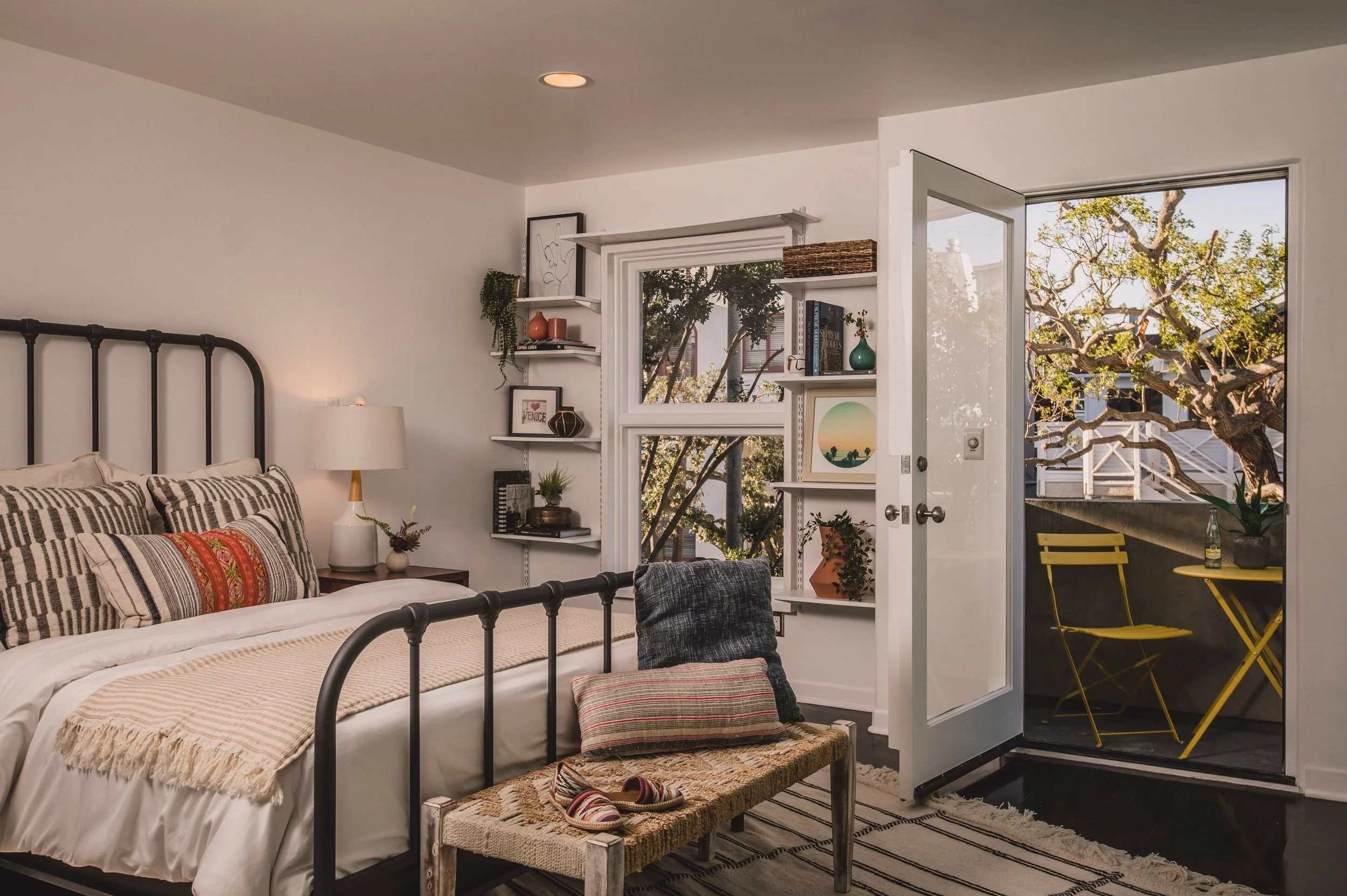 A cozy bedroom with a black metal bed, patterned pillows, a side table with a lamp, and a white bookshelf near an open door leading to a balcony with yellow chairs, and trees outside.