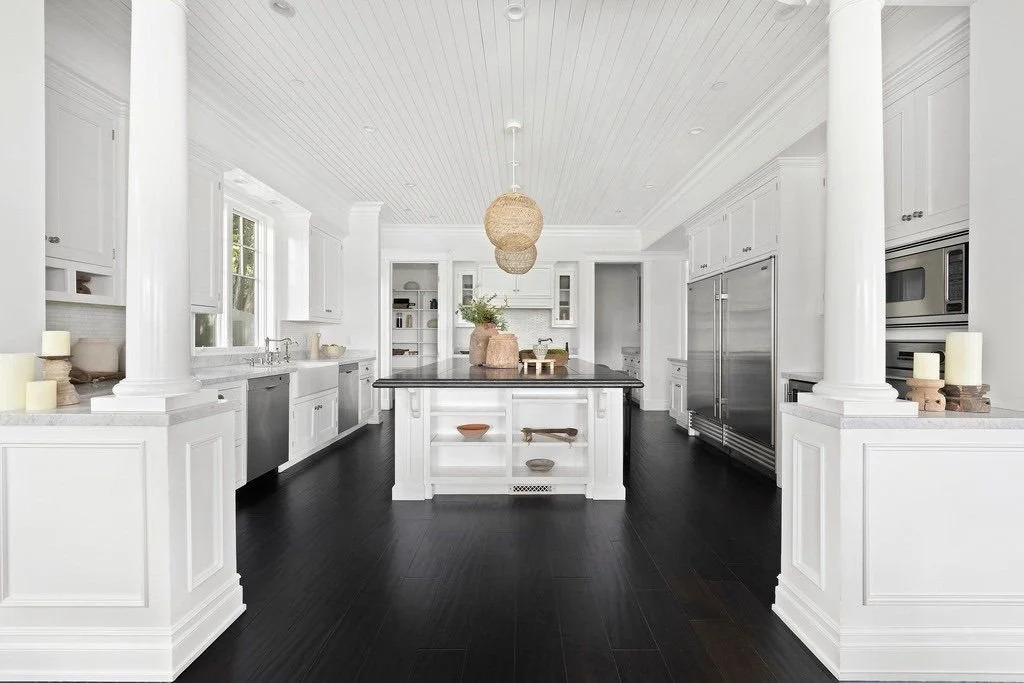 Modern white kitchen with black flooring, large island, pendant lights, and stainless steel appliances.
