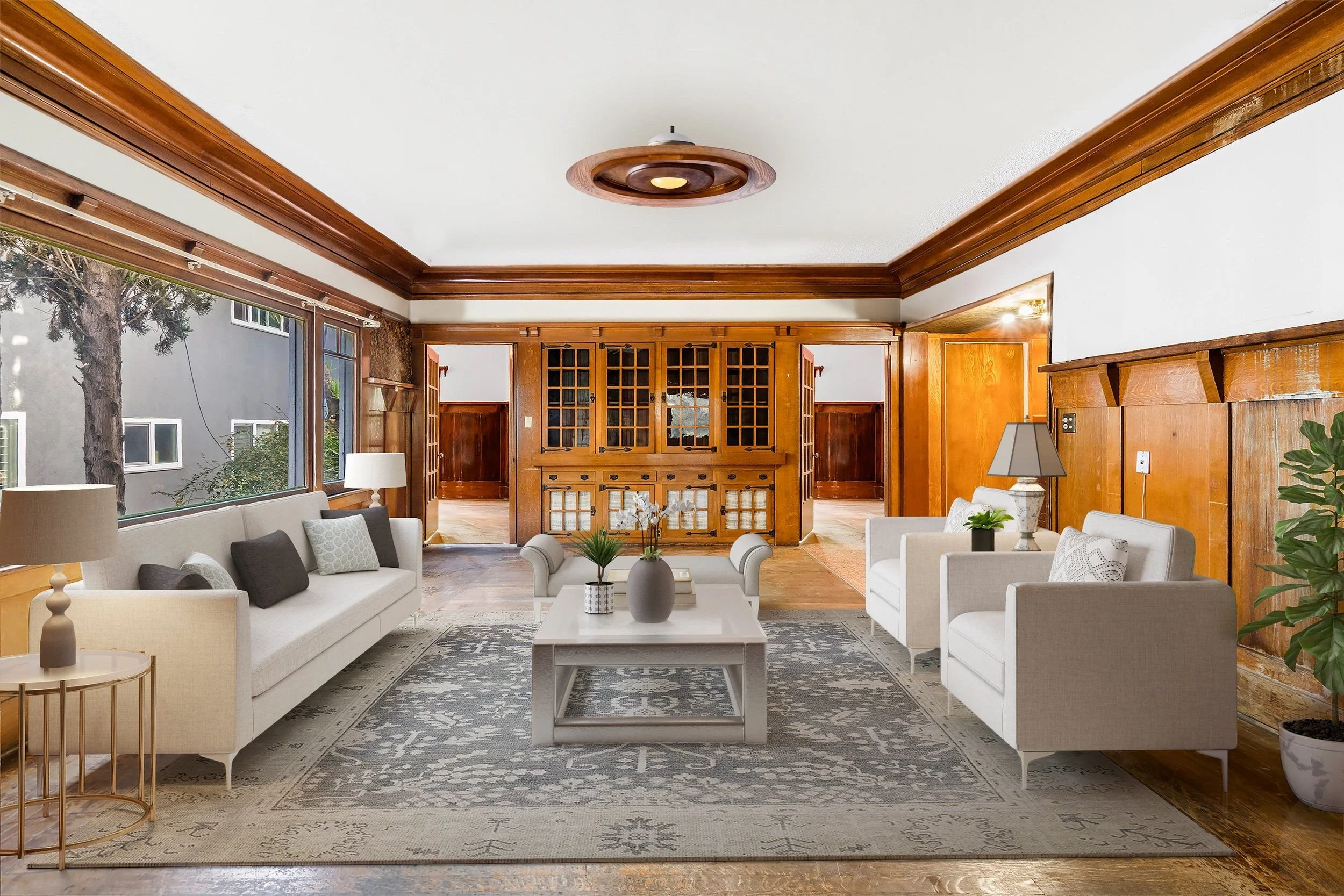 A cozy living room with white sofas, a wooden fireplace with glass doors, large windows showing trees outside, and wooden paneling on walls and ceiling with decorative crown molding.