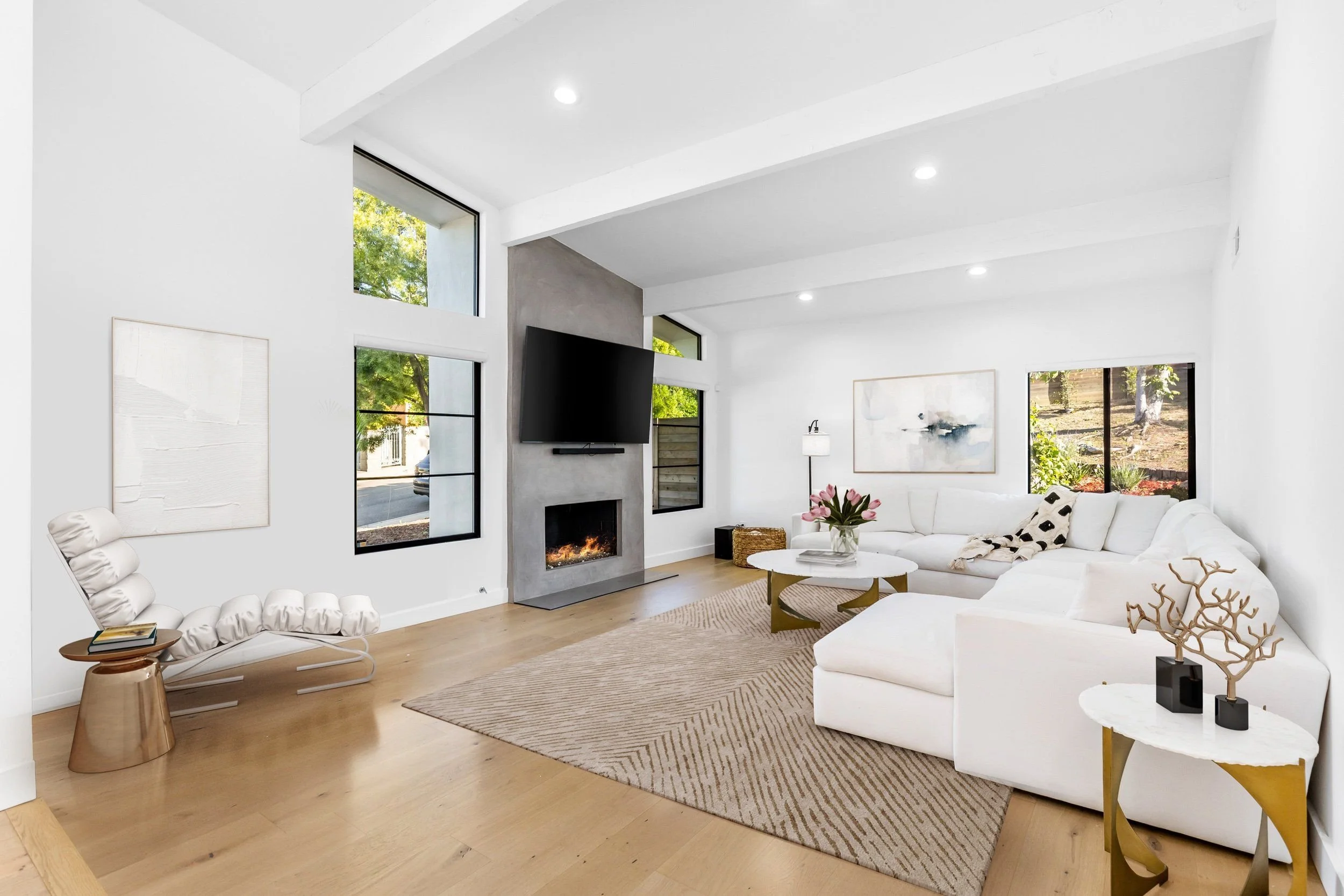 Modern living room with white walls, large windows, a white sectional sofa, a fireplace, and artwork, decorated with gold and white accents, wooden flooring, and ceiling spotlights.