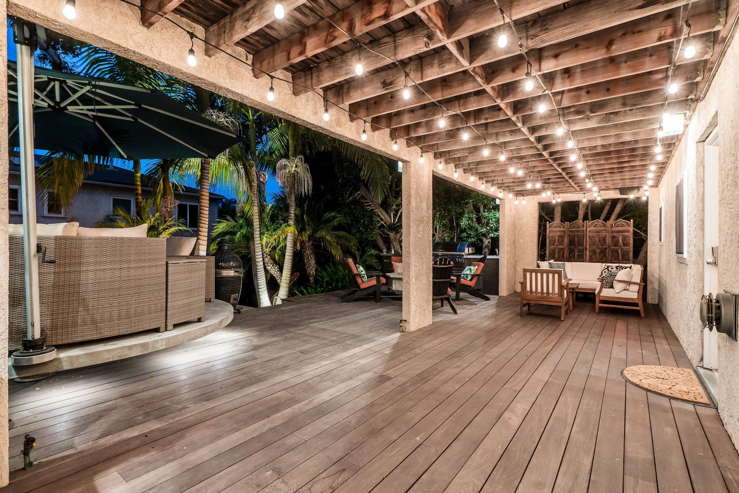 Covered outdoor patio area with string lights hanging from a wooden ceiling and various outdoor furniture, including chairs and a cushioned seating area, with lush trees and plants in the background.