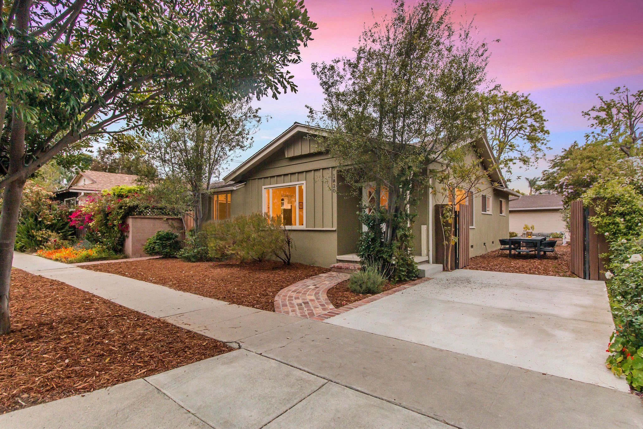 A suburban house with large front windows and a small porch, surrounded by trees and shrubs. The sky shows a colorful sunset with pink and purple hues, and the front yard includes a curved brick pathway and a patio area with outdoor furniture.