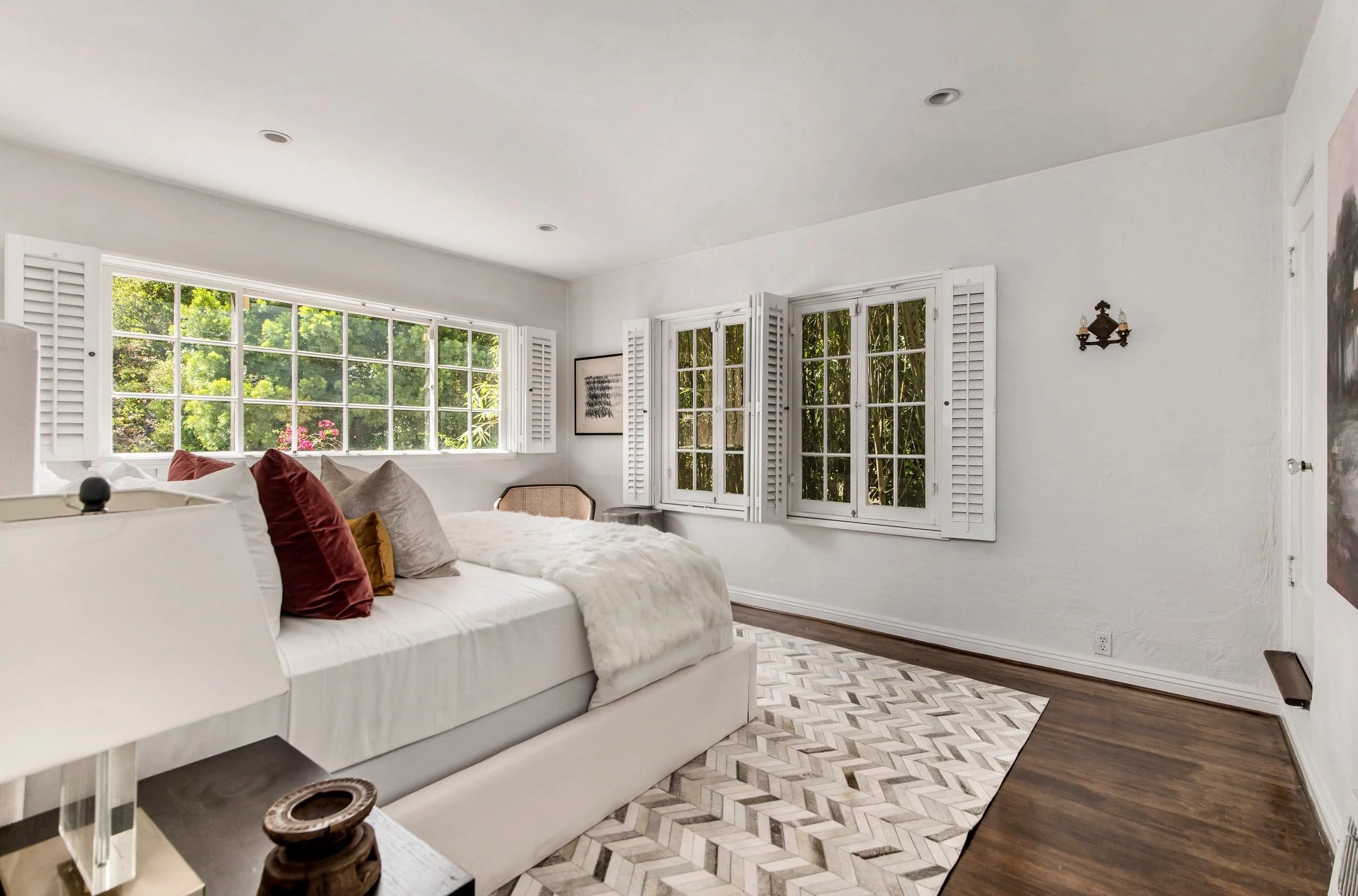 A bright bedroom with large windows and white shutters, white walls, hardwood and patterned tile flooring, a white bed with decorative pillows and a fur throw, a side table with a decorative object, and framed artwork on the wall.