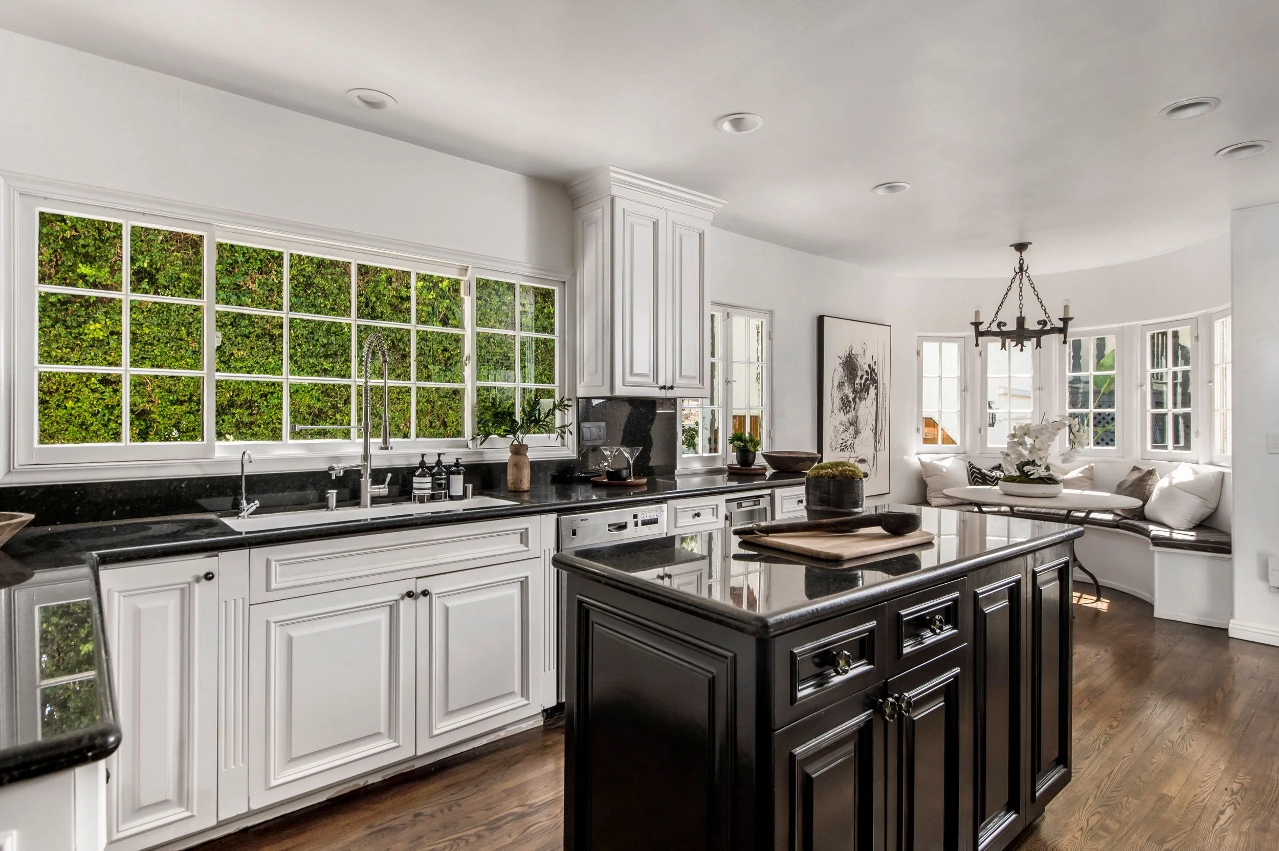 A bright kitchen with large windows, white cabinetry, black granite countertops, and a black island. There are various plants and decor on the counters, and a dining area with a white curved bench and a round table with white flowers. A chandelier ha