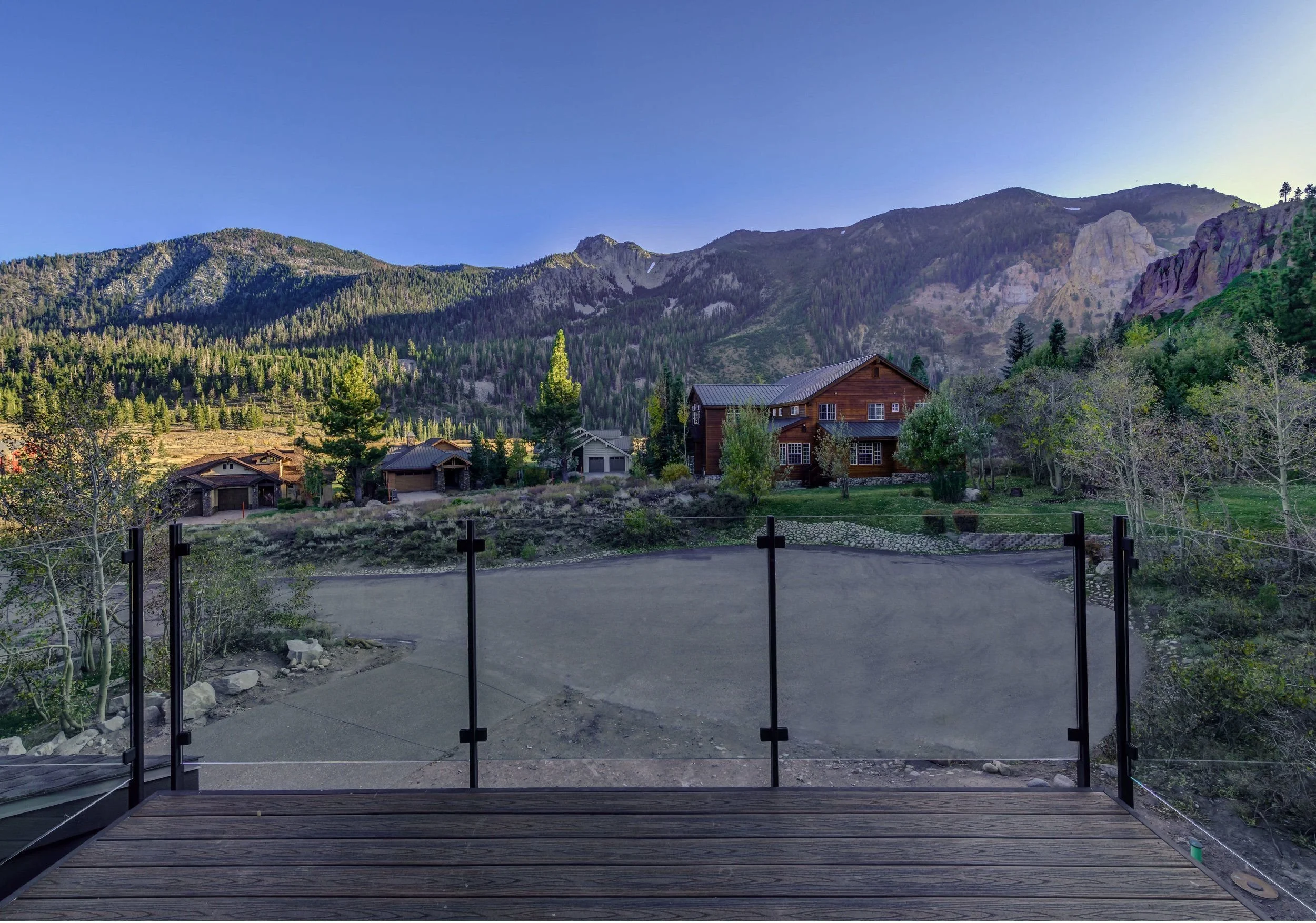 A view of a mountain landscape with green trees, rocky cliffs, and a clear blue sky, viewed from a wooden deck enclosed with glass panels and a black metal railing.
