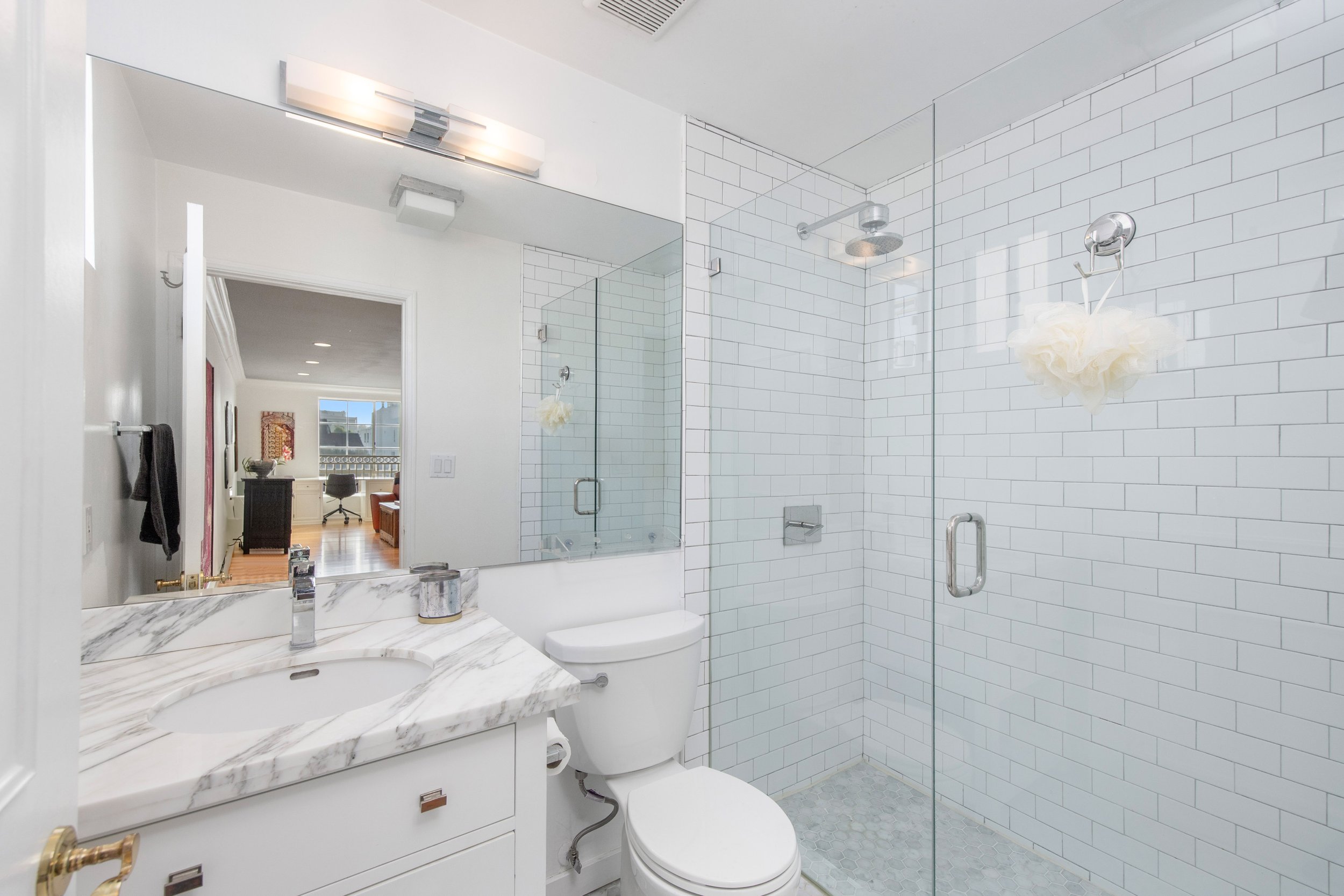 Modern bathroom with white subway tile walls, a glass shower enclosure, a marble vanity with a sink, a large mirror, a toilet, and a nearby doorway leading to a room with wooden flooring and a window.