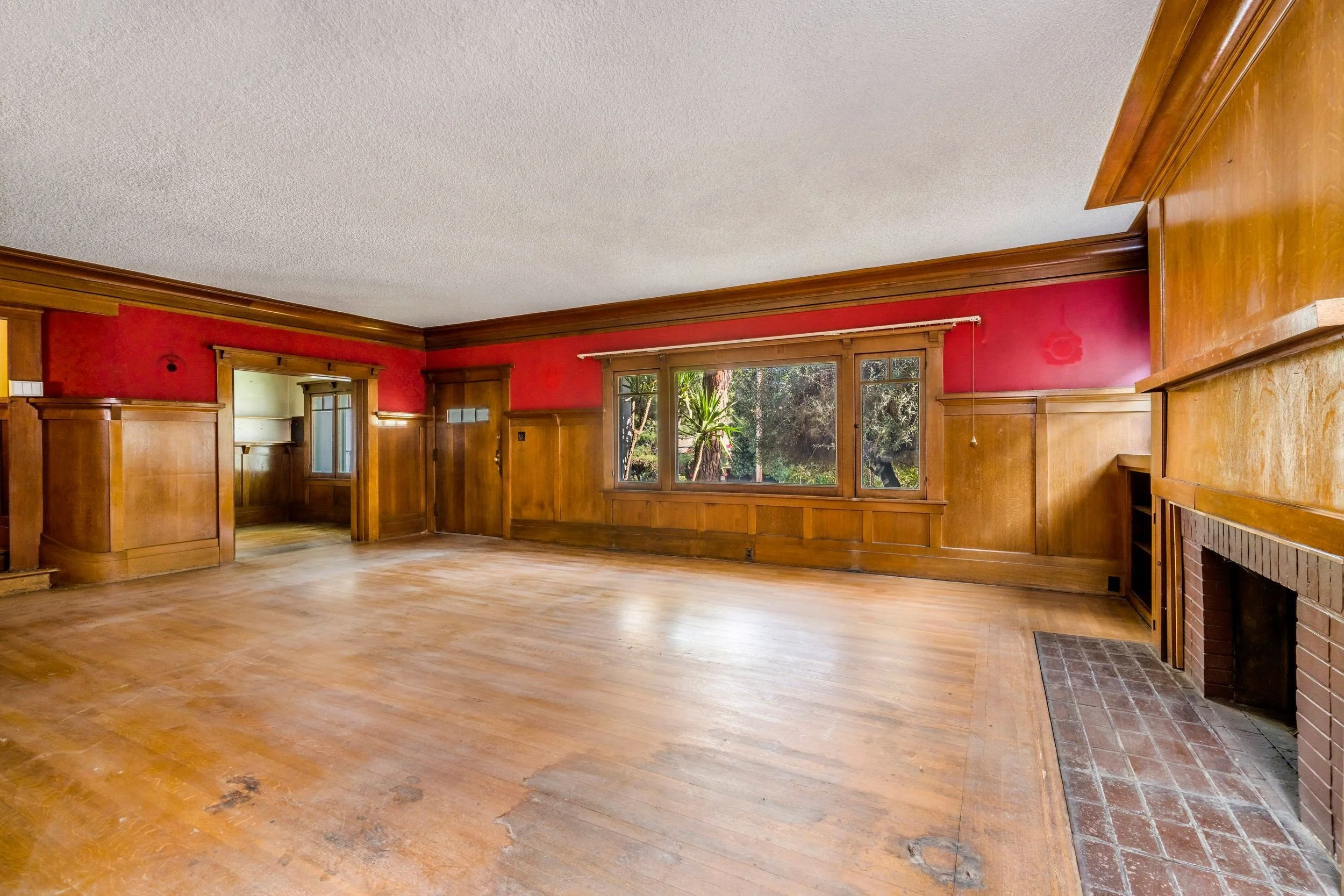 Empty room with hardwood floors, wood-paneled walls, a stone fireplace, and large windows showing greenery outside.