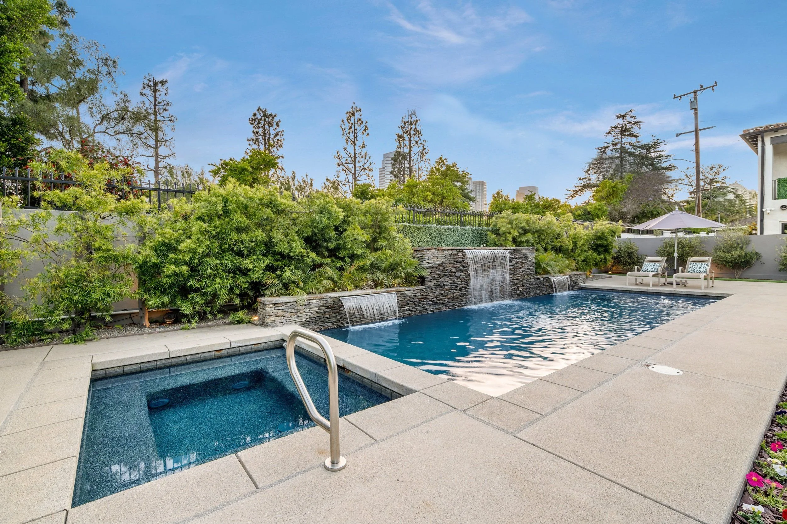An outdoor swimming pool area with a hot tub in the foreground, a waterfall feature, lounge chairs, and an umbrella, surrounded by greenery and trees, with tall buildings in the background under a blue sky.