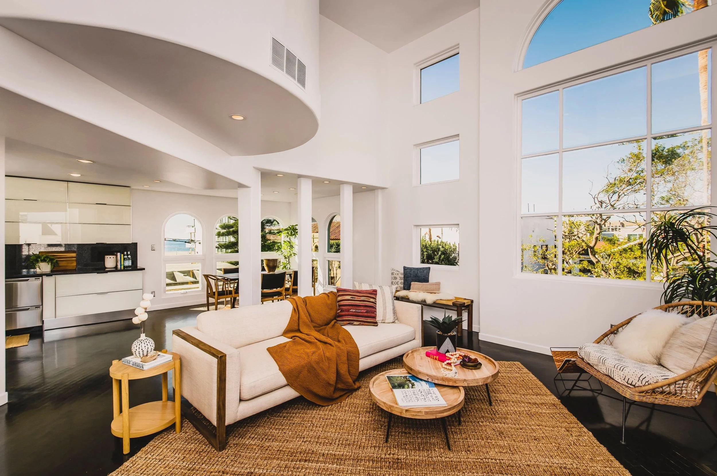 Spacious, modern living room with large high windows letting in natural light, white walls, and decorated with a beige sofa, patterned cushions, and a wooden coffee table with magazines, in a house with a view of trees and blue sky.