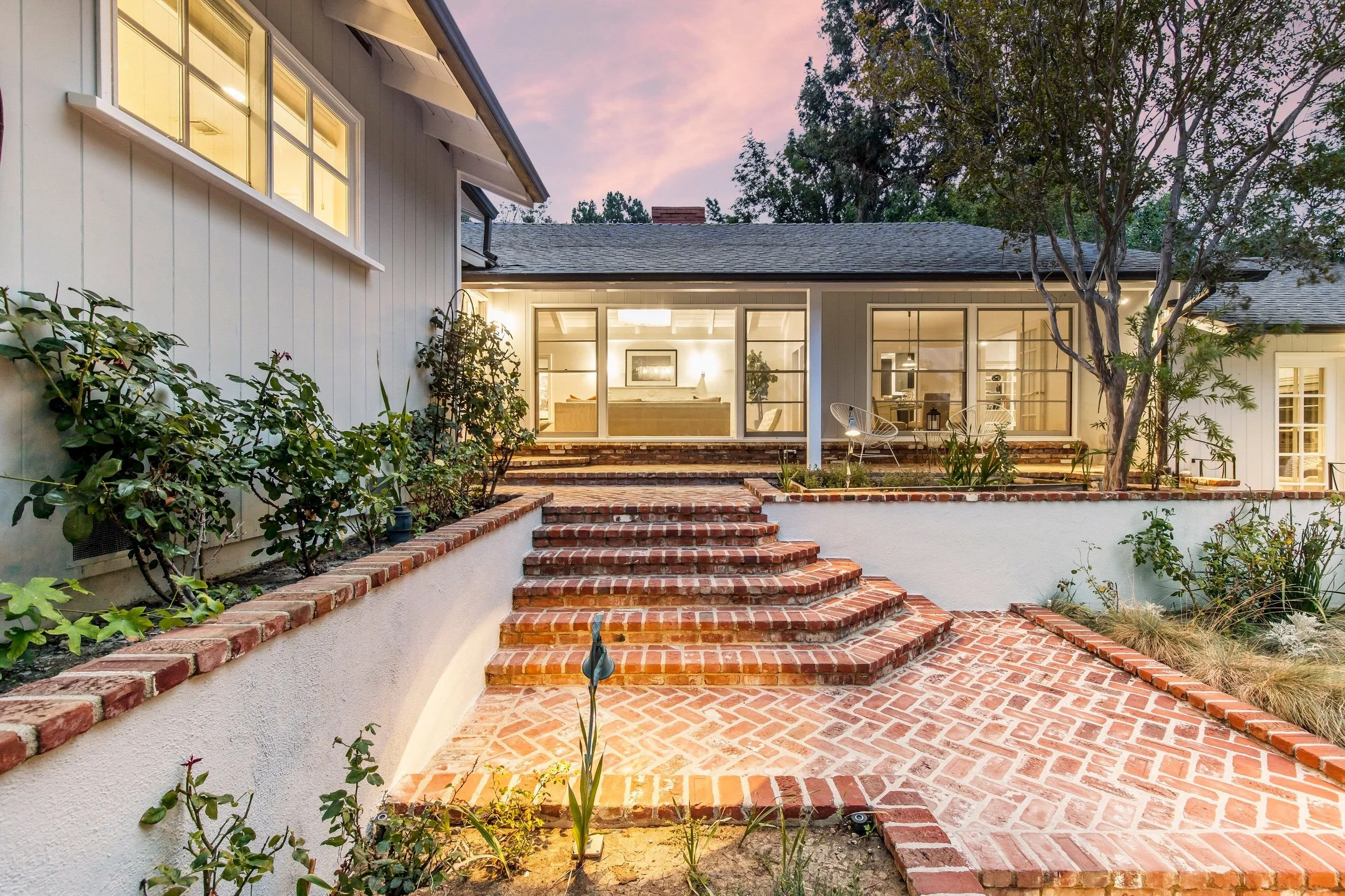 A backyard patio with brick steps and a brick pathway leading to a house with large windows illuminated from inside. There are trees and plants around the patio at sunset.
