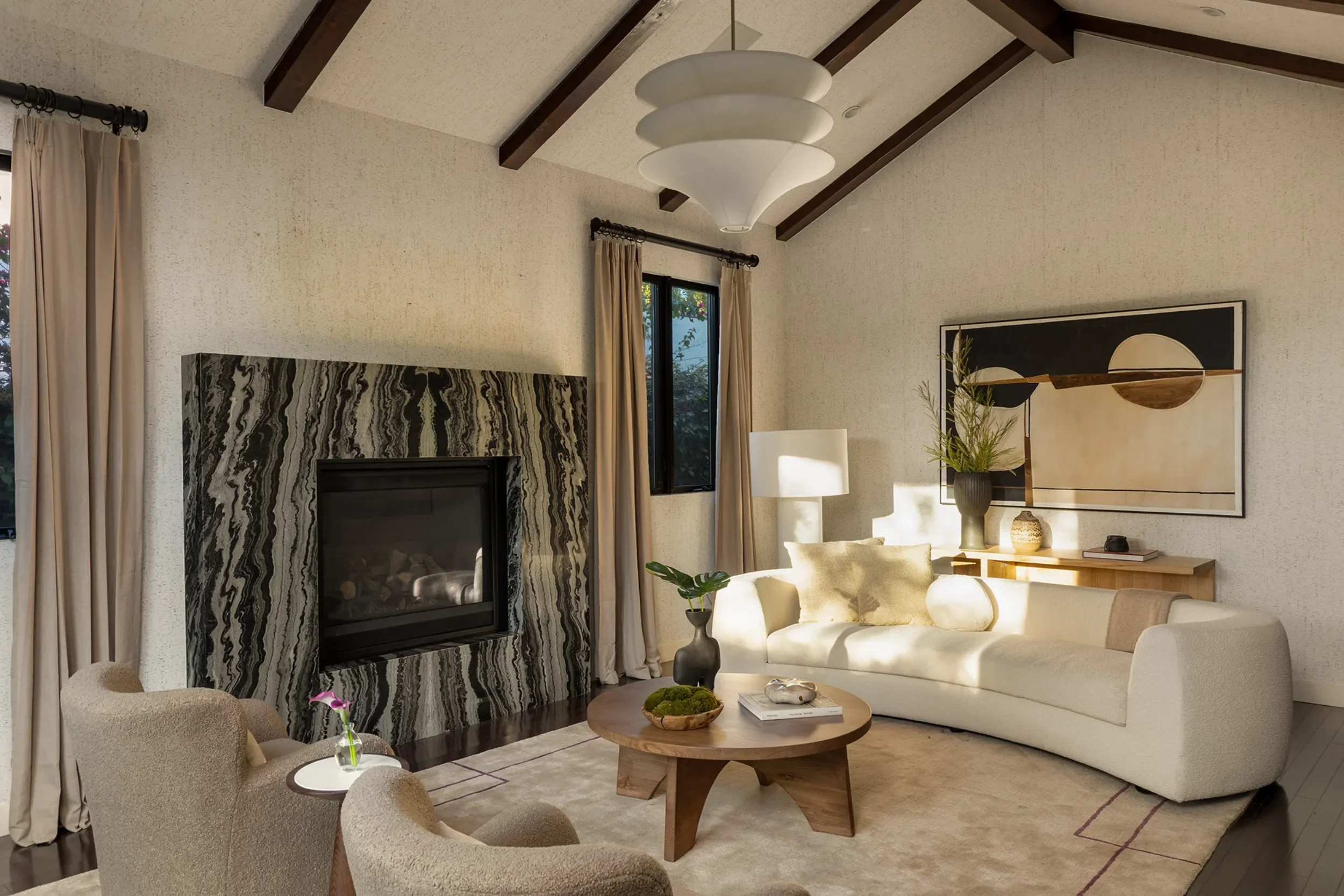 Living room with a white sofa, beige armchairs, a round wooden coffee table, a fireplace with a black and white pattern, a large abstract painting, a black vase with a plant, and a wooden side table with decorative objects. There are beige curtains a